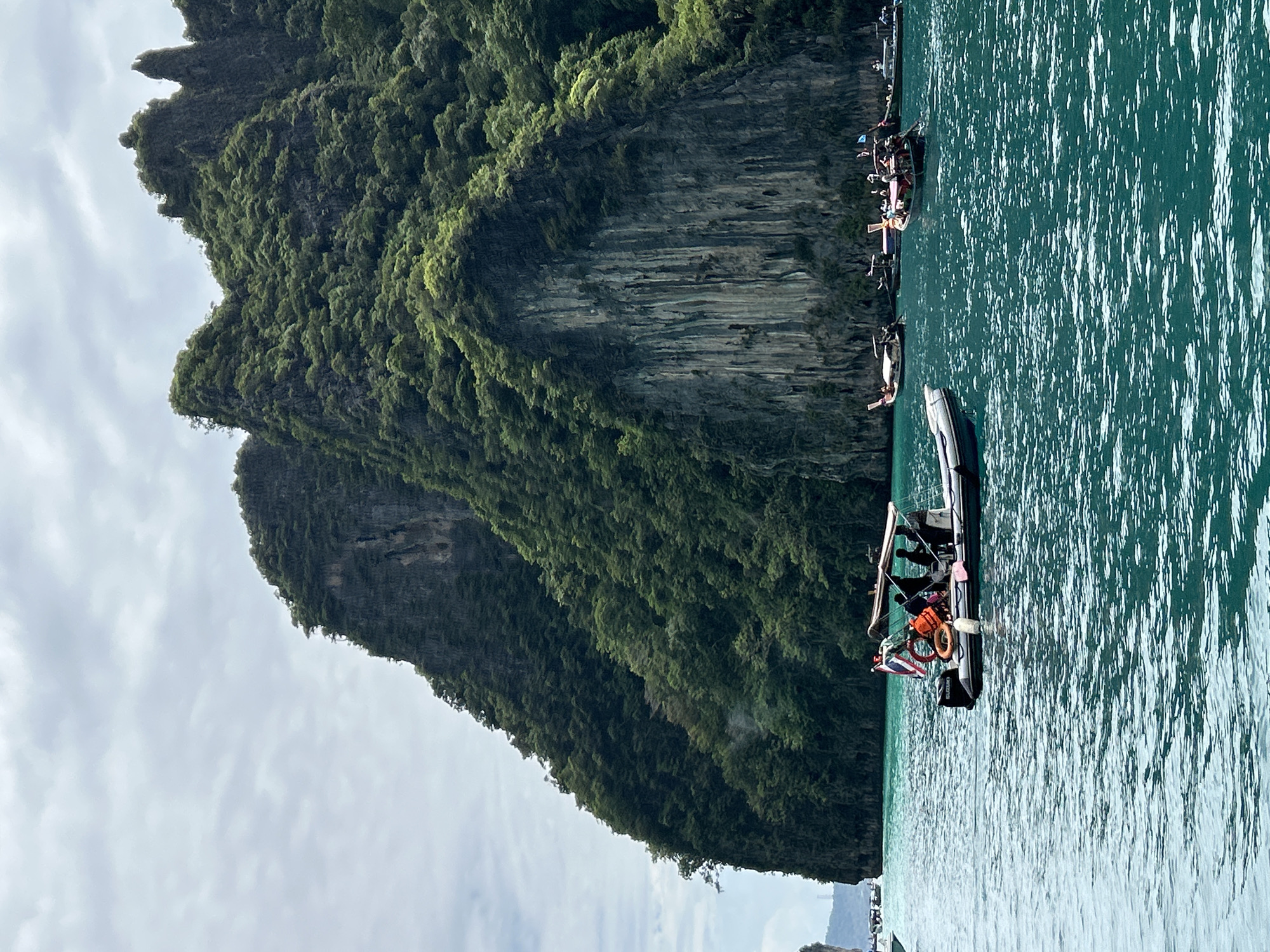 A motorboat carrying at least three people is in the foreground of the image, sailing on turquoise water. The boat has a flag and several life preservers on board. In the background, a large, lush green cliff face rises out of the water, partially obscured by clouds. Several other boats, including traditional longtail boats, are visible in the water. The scene appears to be in a tropical location, likely Thailand, given the presence of longtail boats and the scenery.