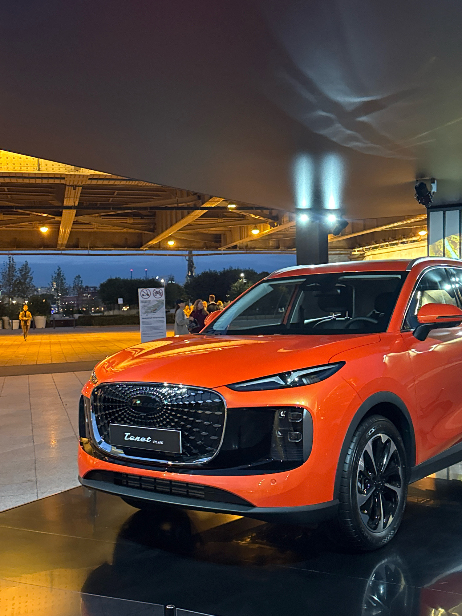 An orange SUV is parked on a dark reflective surface, possibly a display platform, in what appears to be an outdoor urban setting at dusk or night. The car is positioned in the foreground, occupying the right and center of the frame, facing left. It has a prominent, intricately designed grille with a circular emblem and a badge that reads "Tenet PLUS" below it. The car's headlights are on, casting a bright light. Behind the car, a pedestrian walkway is visible, with a few people walking in the middle ground. To the left of the car, a person is seen riding a bicycle. In the background, there are trees, buildings, and a large overhead structure, possibly a bridge or an overpass, with illuminated supports. The sky is a deep blue, indicating it is late evening. The overall scene suggests an exhibition or a public display of the vehicle within a city environment. The location is identified as Moscow, Russia.