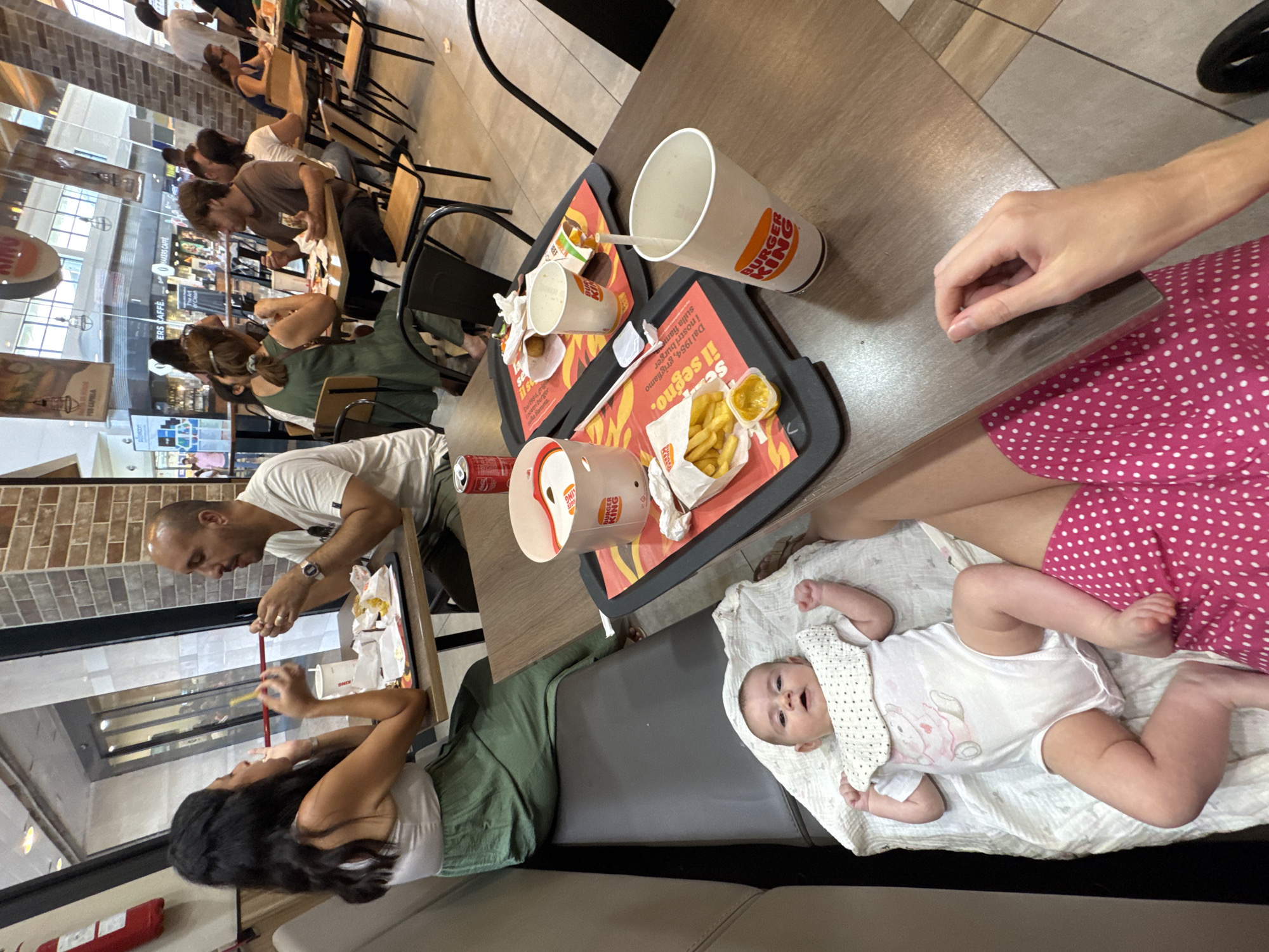 The image shows a family eating at a Burger King restaurant in Carini, Italy. A man and a woman are seated at a table, eating french fries and drinking from Burger King cups. A baby is lying on a blanket on the floor in front of them, wearing a white onesie and a bib. In the background, other diners can be seen eating at tables. The restaurant is brightly lit and decorated with Burger King branding.