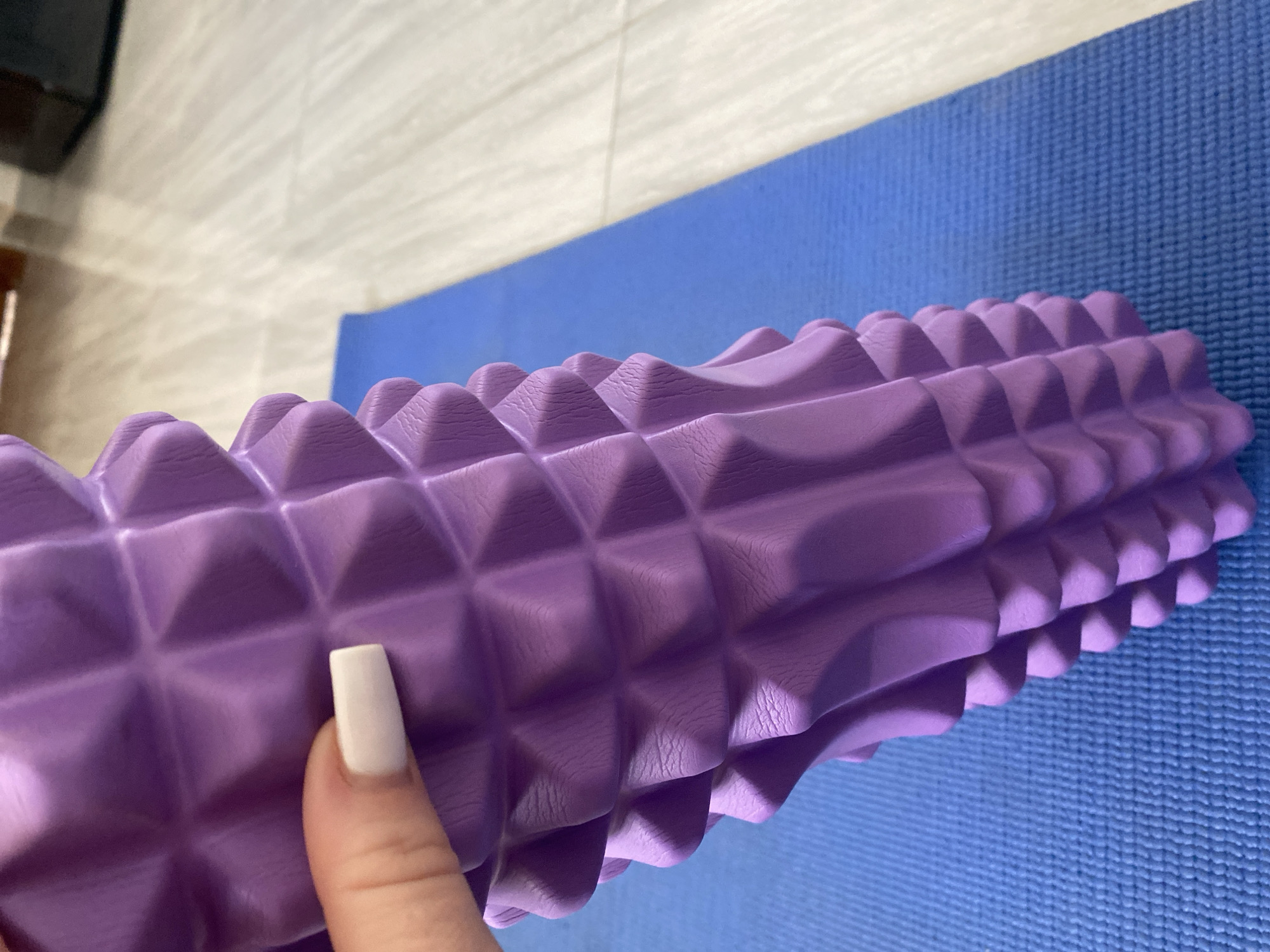 A close-up shot shows a hand with a white-painted fingernail holding a textured purple foam roller. The foam roller is positioned on a blue yoga mat, which is placed on a light-colored wooden floor. The background is blurred, showing hints of furniture and possibly a wall. The scene appears to be indoors, likely in a home or fitness studio. There is no indication of the location being in Sharm El Sheikh, Egypt.