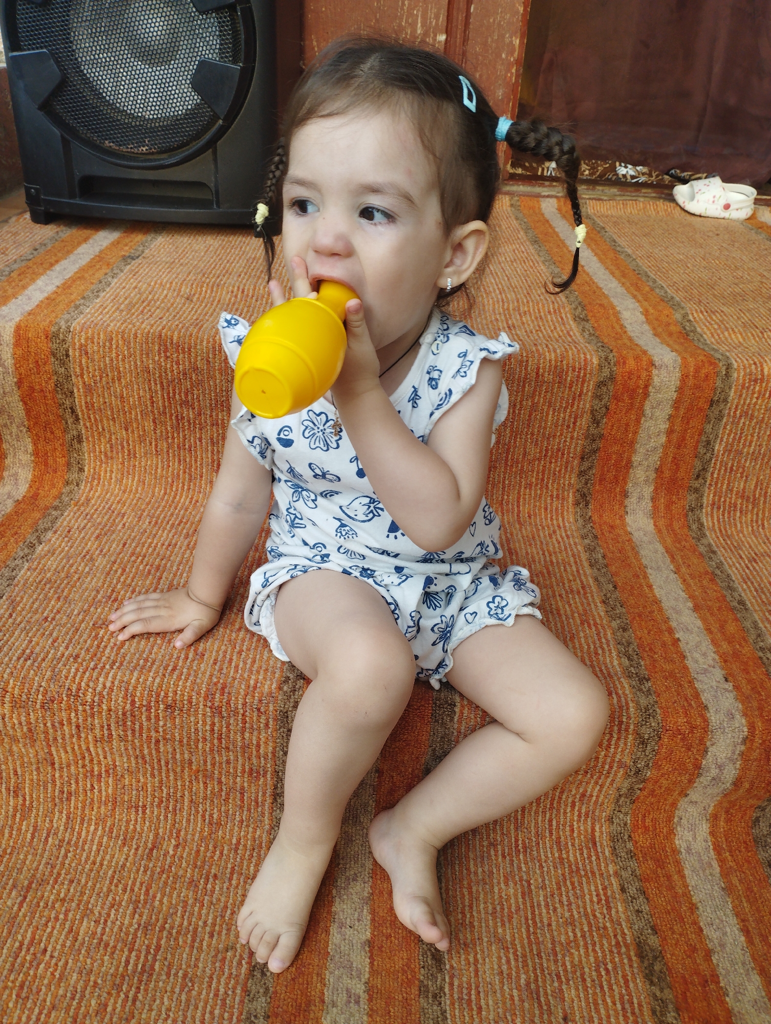 A young girl with dark hair in two braided pigtails is sitting on a striped orange and beige carpeted surface. She is wearing a white romper with blue floral patterns. In her hands, she holds a yellow plastic object, which she is placing into her mouth. To her left, a black speaker is visible. The background appears to be a wooden structure and a brown cloth. A white clog with pink designs is on the floor in the background to the right. The lighting suggests an outdoor or semi-outdoor setting. The overall impression is of a casual, domestic scene. The image was taken in Fălești, Moldova.
