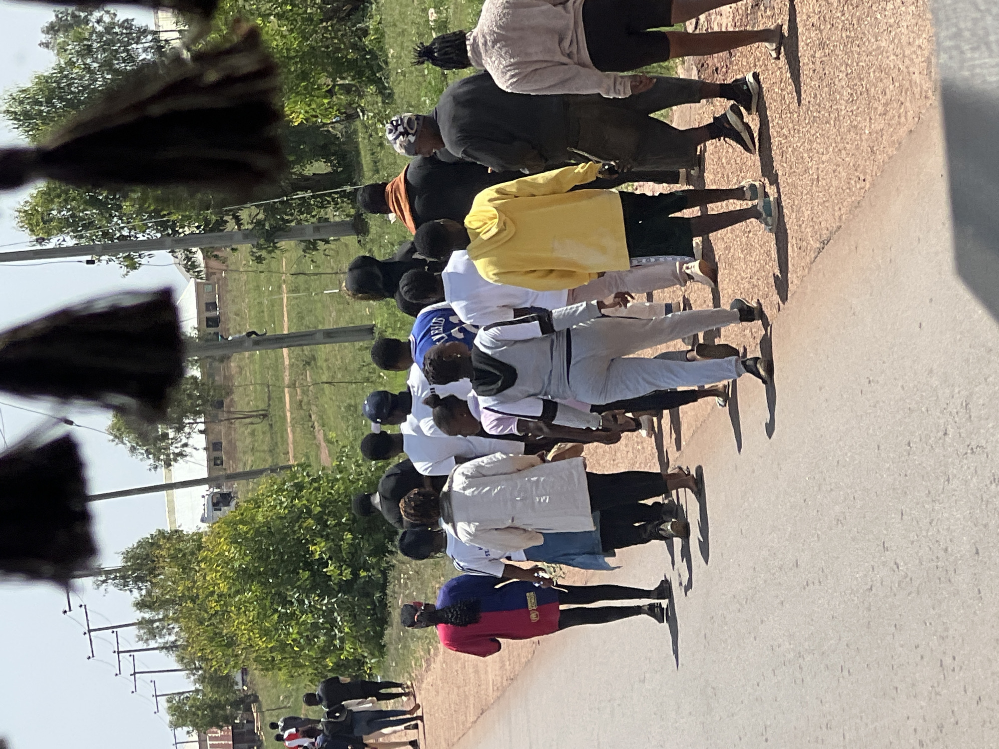 A group of approximately 15 people is walking along a paved road. The people are dressed in casual clothing, including t-shirts, shorts, pants, hoodies and hats. The setting appears to be an outdoor location in Durumi, Nigeria. Trees and buildings are visible in the background. Power lines run along the side of the road. The overall atmosphere is sunny and bright.