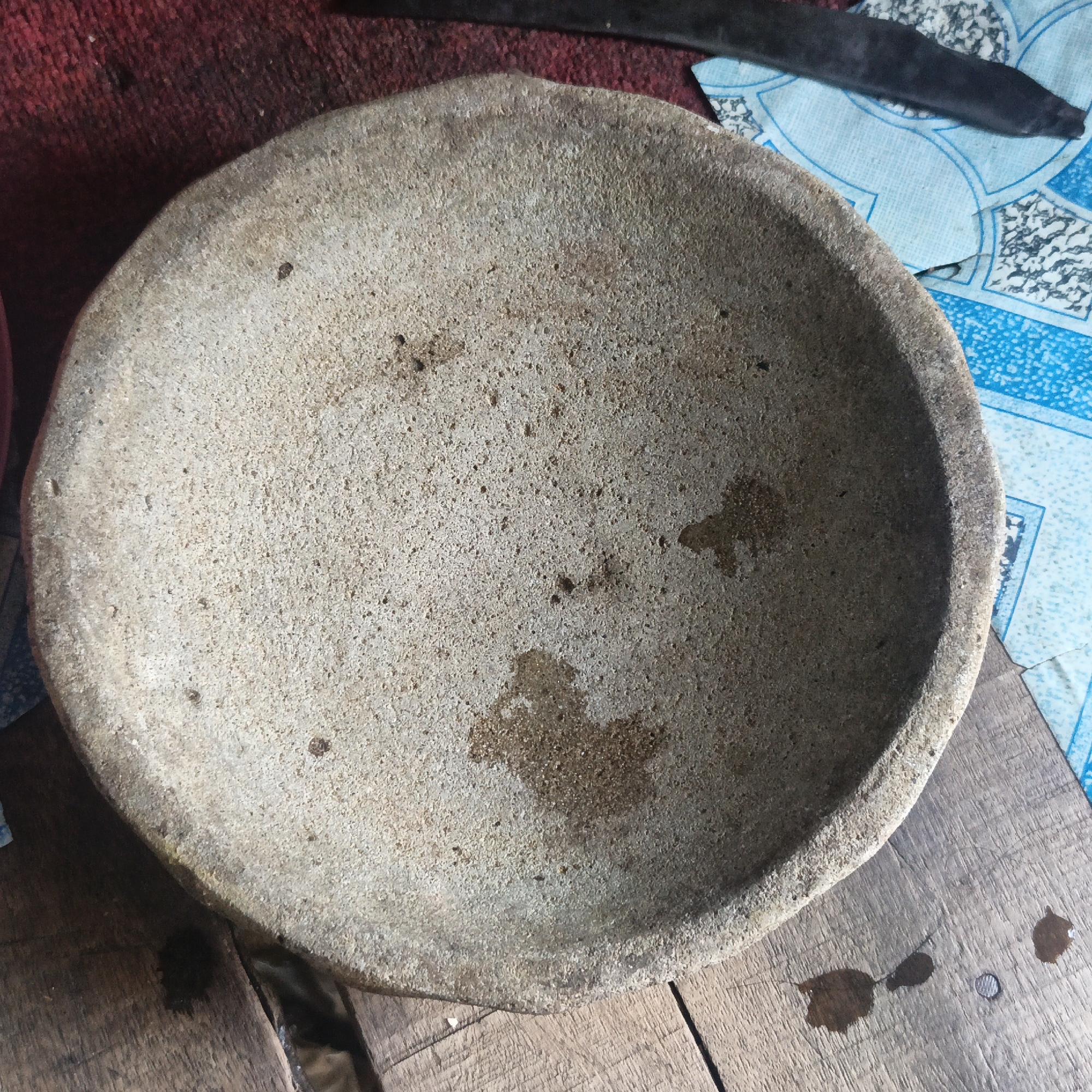 A close-up, top-down view captures a rough, circular stone mortar with a concave interior, resting on a weathered wooden surface. The mortar shows signs of use, with dark, wet patches indicating residue. Surrounding the mortar, a red, textured fabric lies to the left, and to the right, a blue and white patterned plastic sheet is visible, partially obscured by a dark, elongated object that resembles a pestle or blade. The scene appears to be in an outdoor or rustic setting in Sungai Rasau, Indonesia.