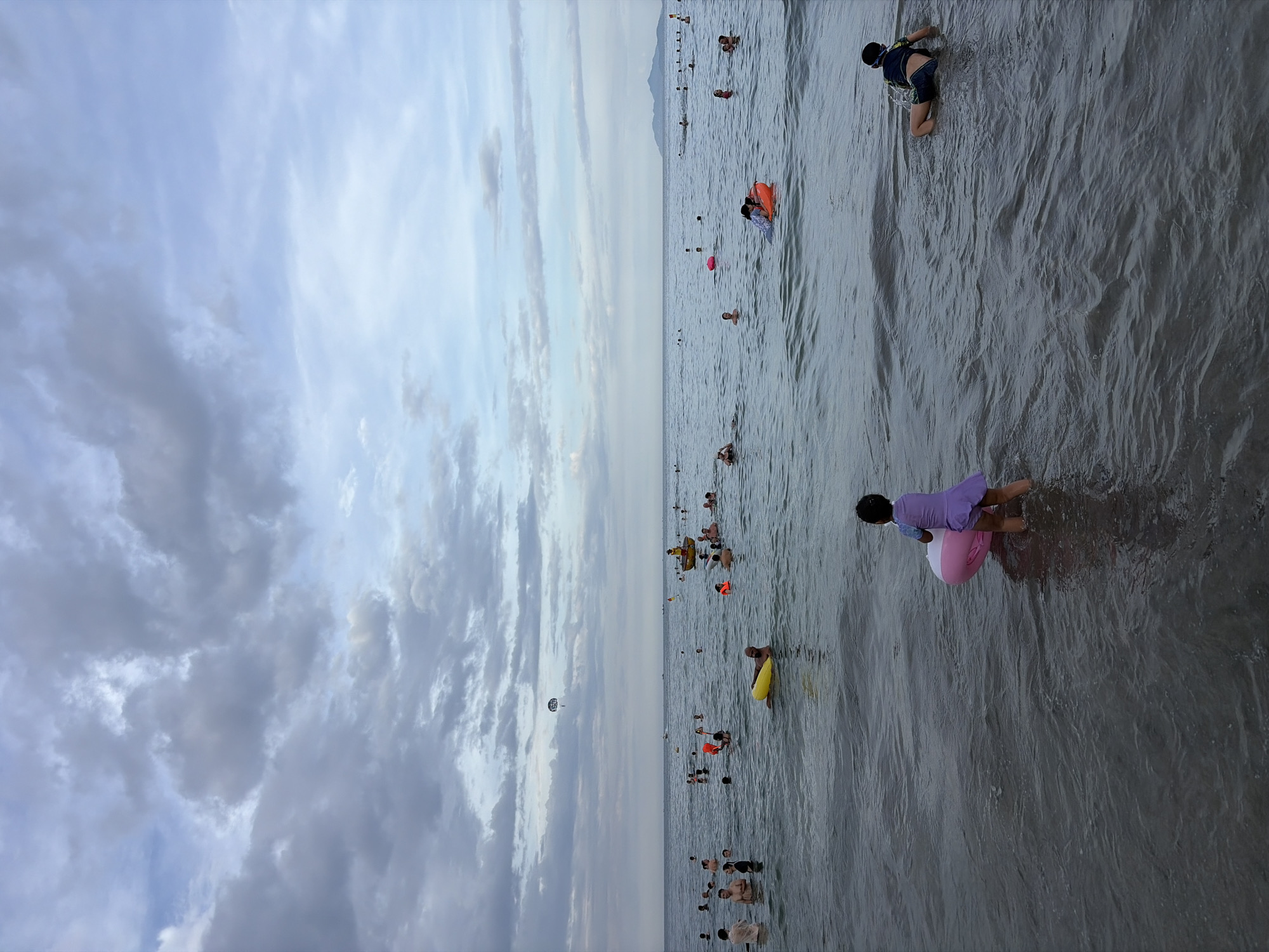 The image depicts a crowded beach scene with numerous people swimming and wading in the ocean under a cloudy sky. In the foreground, a young child in a purple swimsuit stands in shallow water with a pink and white inflatable ring. Further out in the water, many individuals are visible, some with inflatable rings or flotation devices. A yellow rescue boat with a person in it is positioned in the middle ground. In the distance, a parasail can be seen in the sky above the water, and a hazy coastline with mountains is visible on the horizon. The overall atmosphere suggests a recreational day at the beach. This scene is likely on one of the beaches in Đà Nẵng, Vietnam, such as Mỹ Khê Beach or Phạm Văn Đồng Beach, which are known for their popularity and extensive shoreline.