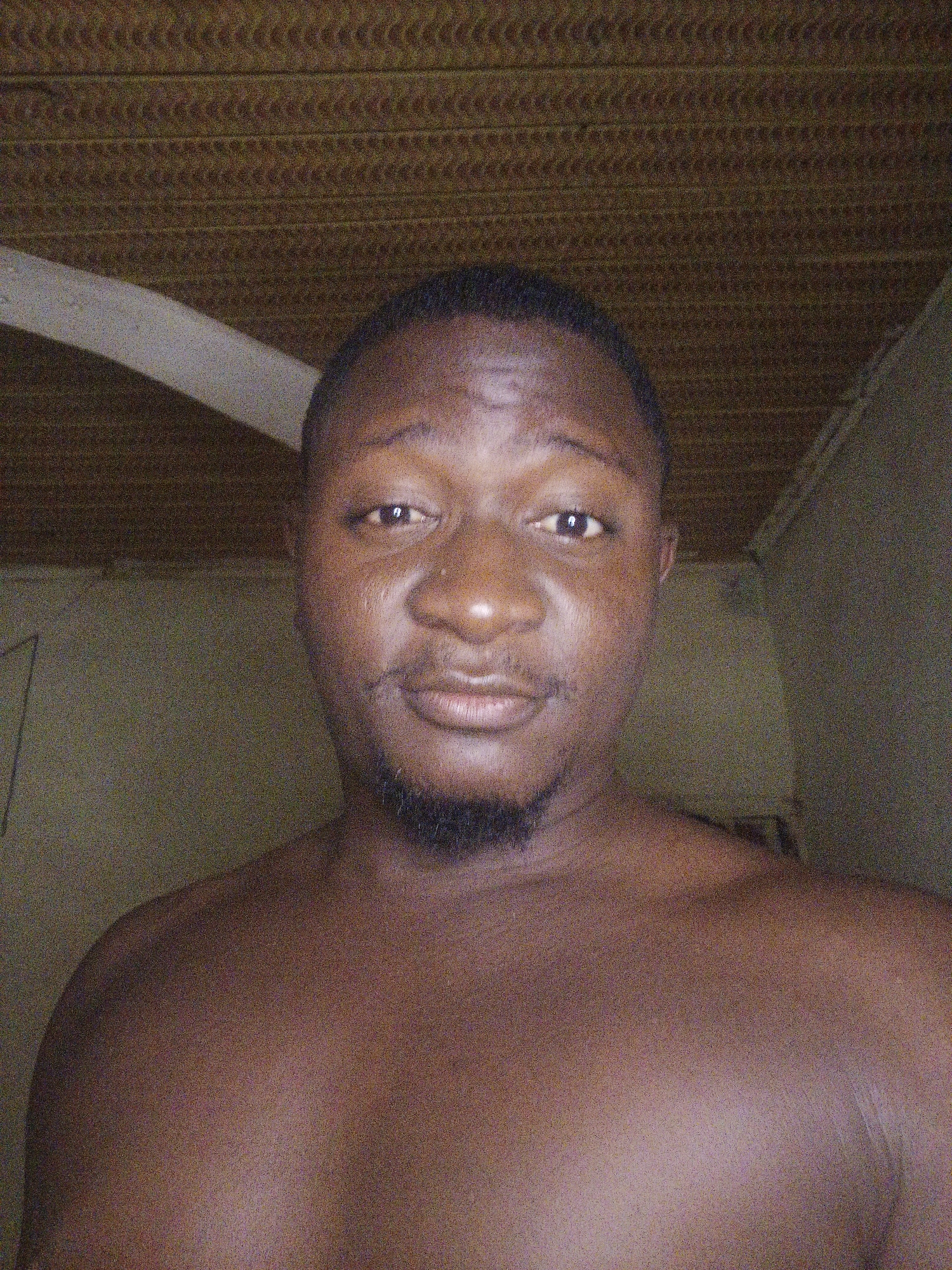The image is a close-up, head-and-shoulders selfie of a shirtless Black man with dark skin and a short haircut. He has a light beard and is looking directly at the camera with a slight smile. The background appears to be an indoor setting with a textured ceiling and a wall that is partially obscured. There is a light-colored diagonal element on the left side of the frame, possibly part of a structure or piece of furniture. The lighting is somewhat dim, and the image has a grainy quality.