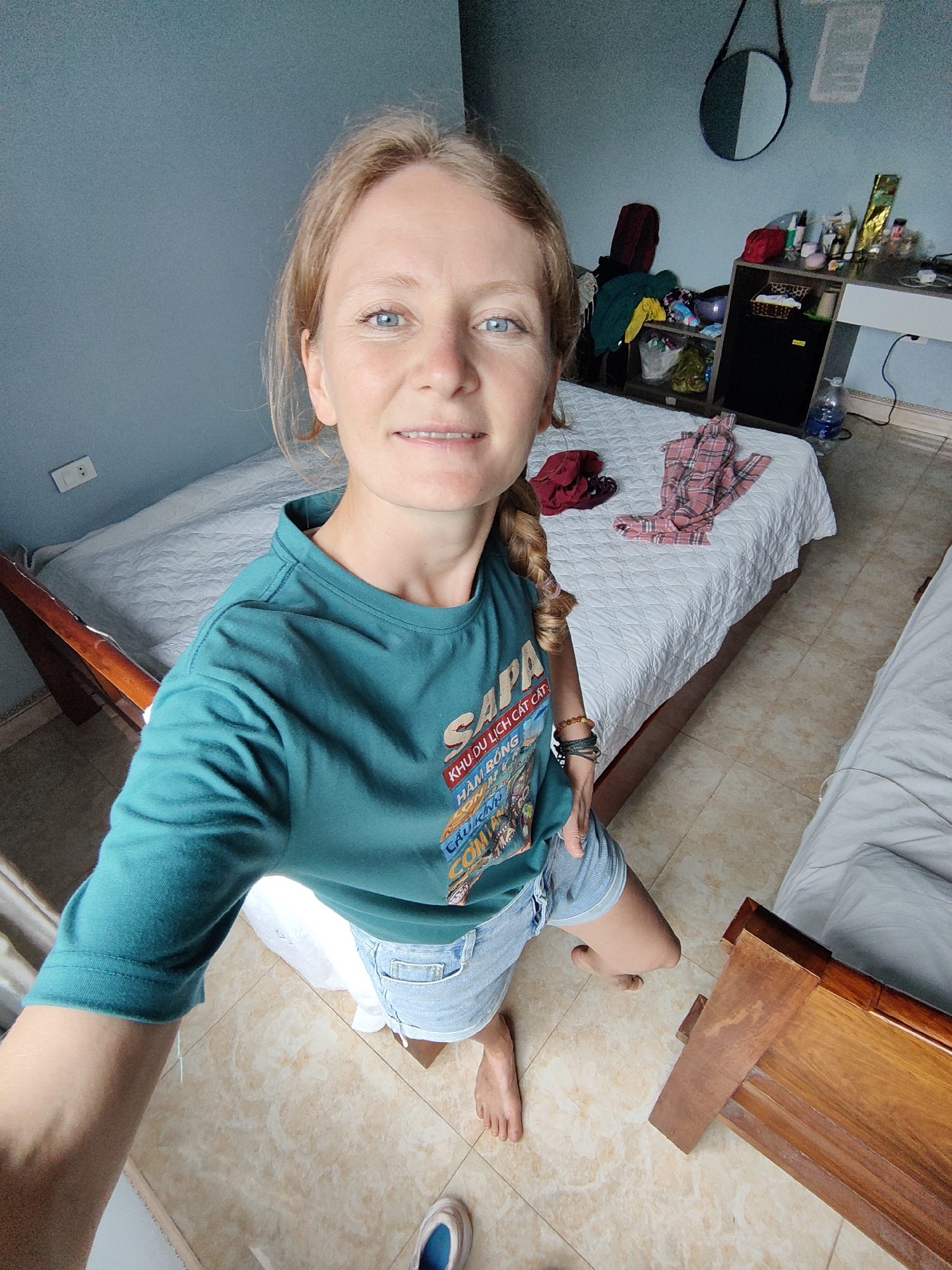 A woman with blonde hair tied in a braid and blue eyes smiles gently while taking a high-angle selfie. She is wearing a teal long-sleeved t-shirt with a colorful graphic design and light blue denim shorts. Her feet are bare, with one light blue and white shoe visible on the tiled floor. Several bracelets adorn her left wrist.
The image captures an indoor room, likely a bedroom or hotel room. The walls are painted a muted blue-grey. Two wooden-framed beds with white quilted bedspreads are visible, one in the foreground and another behind the woman. The bed behind her has a plaid shirt and a dark red garment casually placed on it. Against the far wall, a cluttered dresser holds various toiletries and items, with a black mini-fridge underneath. A round, partially reflective mirror hangs on the wall above the dresser. A large clear water bottle stands on the floor next to the dresser. A white power outlet is visible on the wall near the closest bed. The floor is covered with light brown tiles.
The text on the woman's t-shirt clearly displays "SAPA" at the top, followed by "KHU DU LỊCH CÁT CÁT", "HAM RONG", and "CAU KINH", indicating a souvenir from a Vietnamese tourist destination. The natural light suggests it is daytime. The scene and the visible text are consistent with a traveler's stay in Bai Chay Ward, Vietnam.
