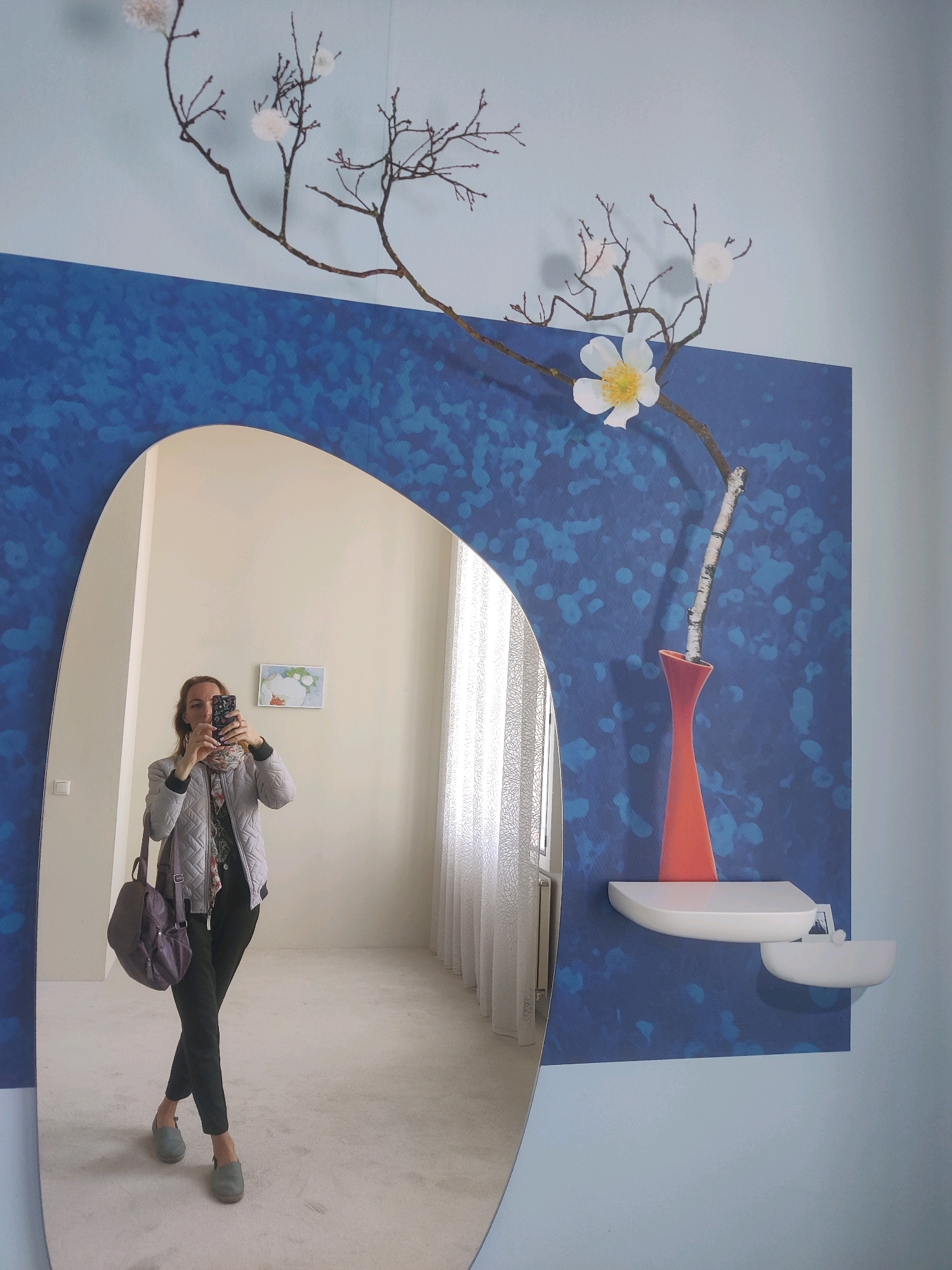 A woman, dressed in a quilted grey jacket, a patterned scarf, and dark trousers, stands in front of a large, oval mirror, taking a selfie with her phone. She is wearing grey clogs and carrying a purple backpack on her left shoulder. The mirror reflects a room with cream-colored walls and a textured white carpet. A small framed picture with a white flower motif hangs on the wall behind her.
The mirror is framed by a blue wall covering with a speckled pattern. On this blue wall, a decorative branch with white pom-pom-like ornaments and a single white flower with a yellow center is mounted. A thin, birch-like twig is placed in an orange vase on a white shelf. The wall itself is painted a light blue.
The scene appears to be an indoor space, likely a home or a gallery, given the decorative elements. The woman's attire suggests casual or semi-casual wear. The overall impression is that of a staged display or an art installation within a room, with the mirror serving as a central element for interaction and reflection. The location is identified as the city of Winterswijk, Netherlands.