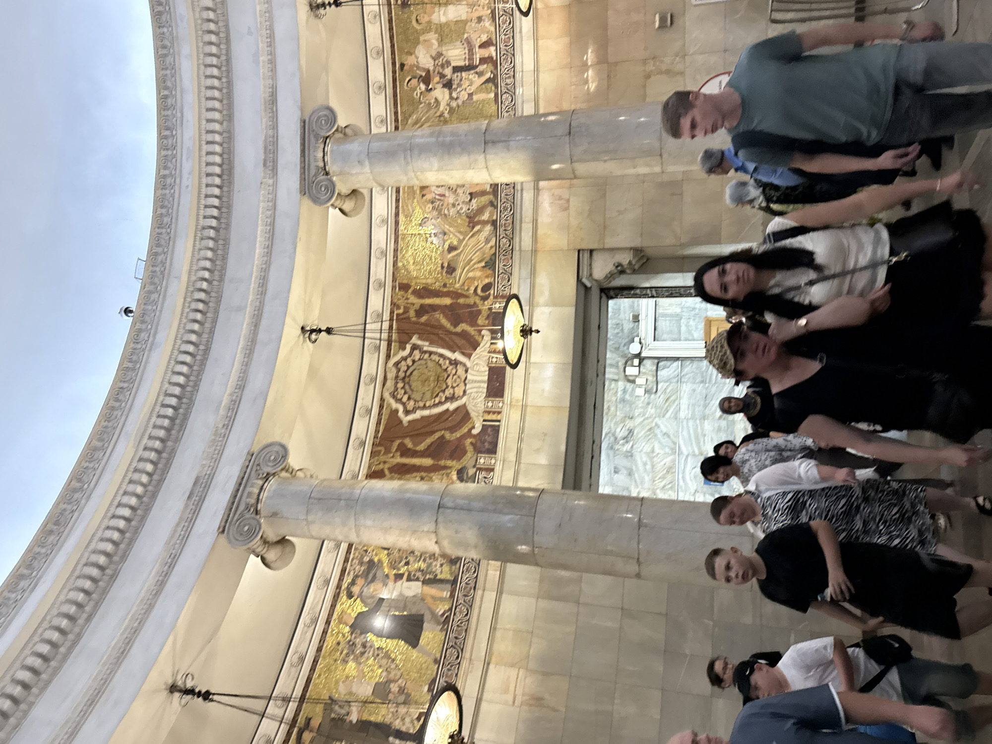 This image depicts a group of people walking through a grand, marble-clad interior. The architecture features large, ornate columns, decorative mosaics depicting figures in traditional clothing, and curved ceilings with intricate moldings. Several pendant lights hang from the ceiling, illuminating the space. The people in the foreground are casually dressed and appear to be moving through the area, some interacting with each other. The background shows an arched doorway leading to another part of the building, suggesting a public space like a metro station or a museum. The overall impression is one of historical grandeur and bustling activity. This location is likely the Mayakovskaya metro station in Moscow, Russia, known for its ornate mosaics and architectural splendor.