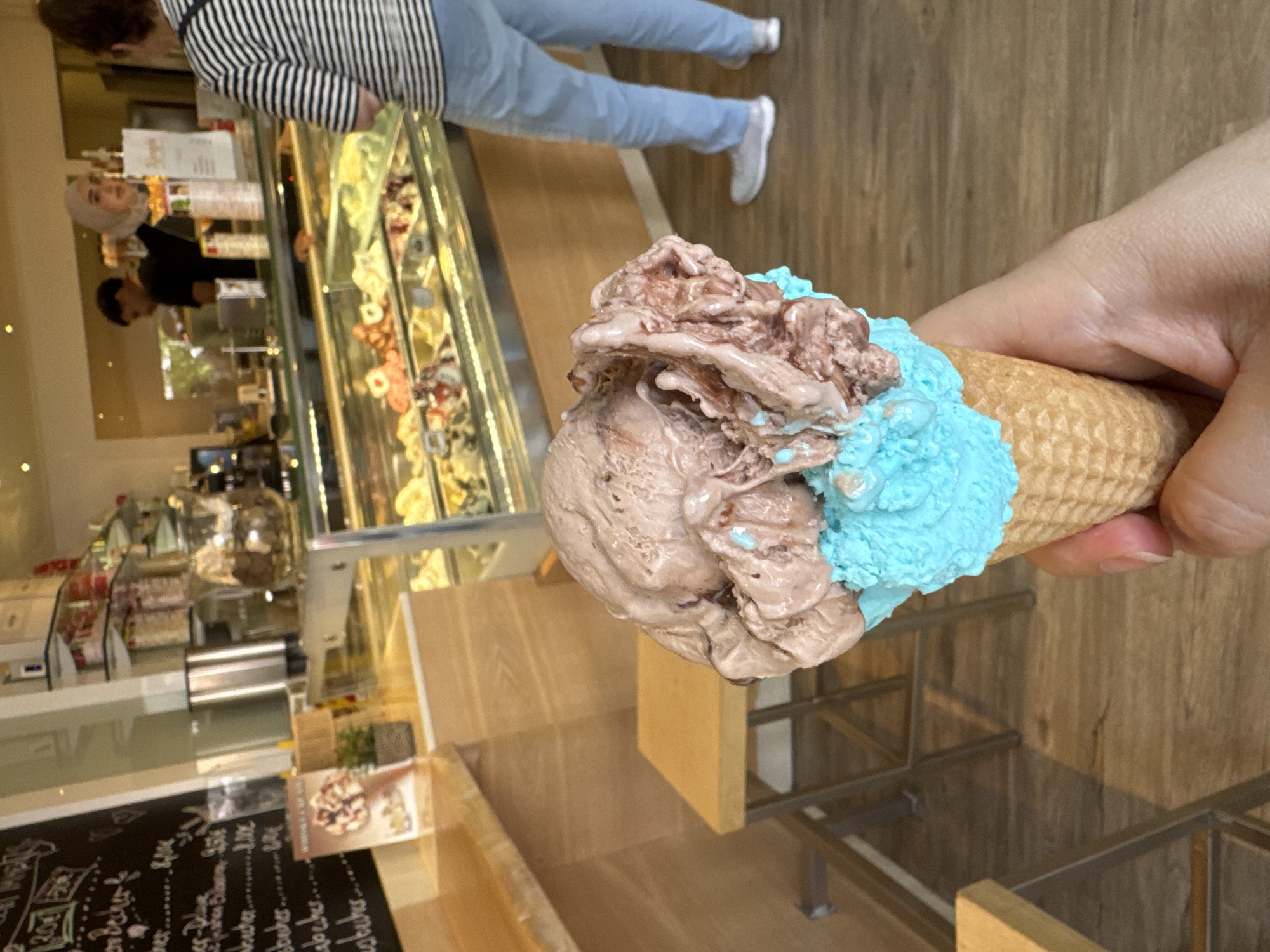 A person's hand holds a two-scoop ice cream cone, with the bottom scoop being bright blue and the top scoop appearing to be chocolate. The ice cream is in the foreground, with a blurred background that appears to be an ice cream shop. In the shop, there are display cases filled with various ice cream flavors, a counter, shelves stocked with goods, and several people. One person, wearing a hijab, is behind the counter, and another person, wearing a striped shirt and light blue pants, is in front of the counter looking towards the display cases. A blackboard menu is visible on the left side of the image with handwritten text. The overall scene suggests a casual, indoor environment, likely a gelateria or ice cream parlor. Based on the menu's handwritten text and likely prices (e.g., "8,00€", "9,50€"), and general store appearance, this location appears to be an ice cream shop in Rheine, Germany.