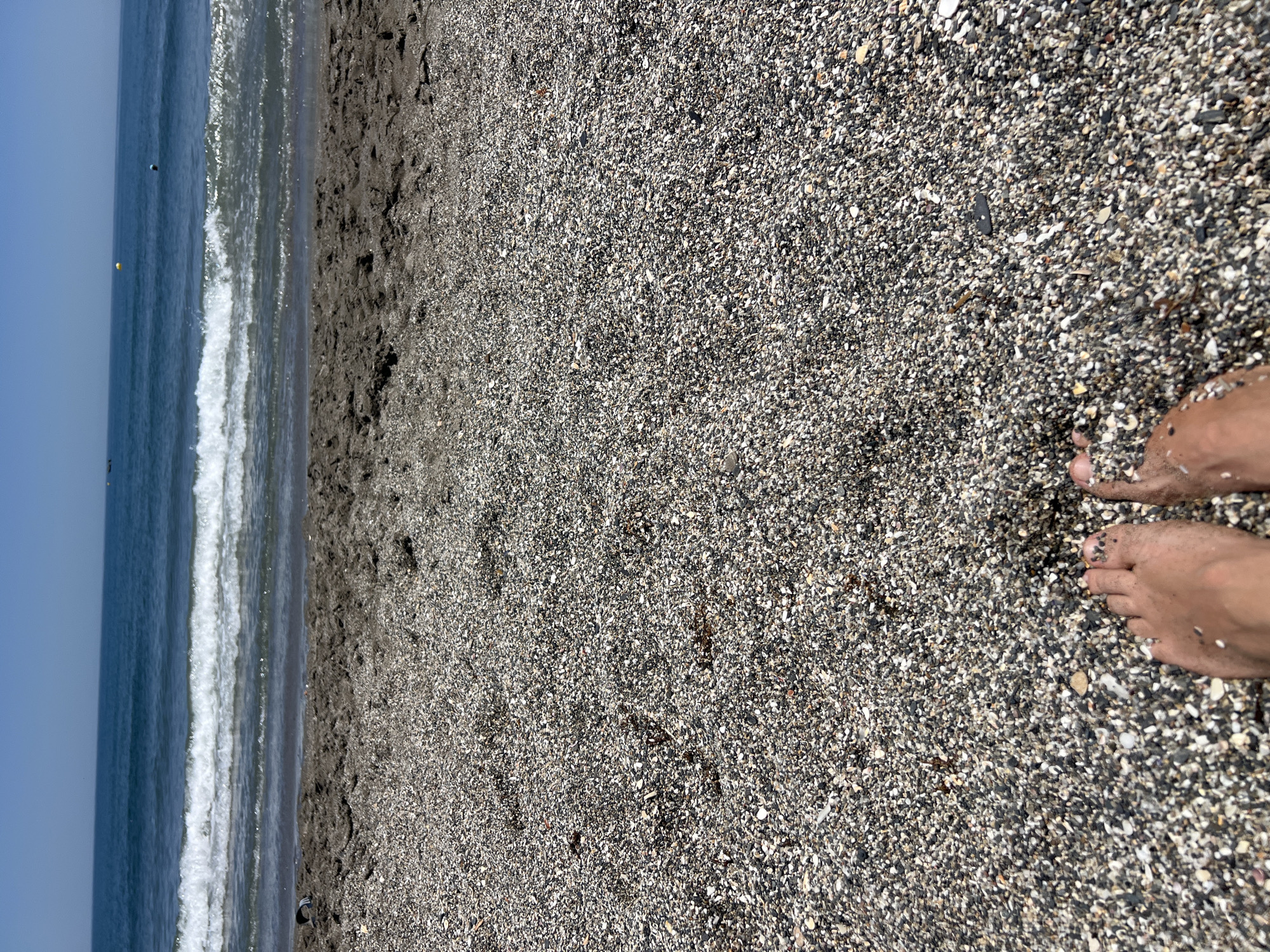 The image captures a perspective of a person's feet resting on a pebble beach, facing the Mediterranean Sea. The sand is a mixture of dark and light pebbles, with some damp patches closer to the water. Gentle waves are breaking on the shore, with a clear blue sky overhead. In the distance, a few buoys are visible in the sea, suggesting a marked area. The scene is peaceful and evokes a sense of relaxation on a sunny day. The location is implied to be a beach in San Roque, Spain.