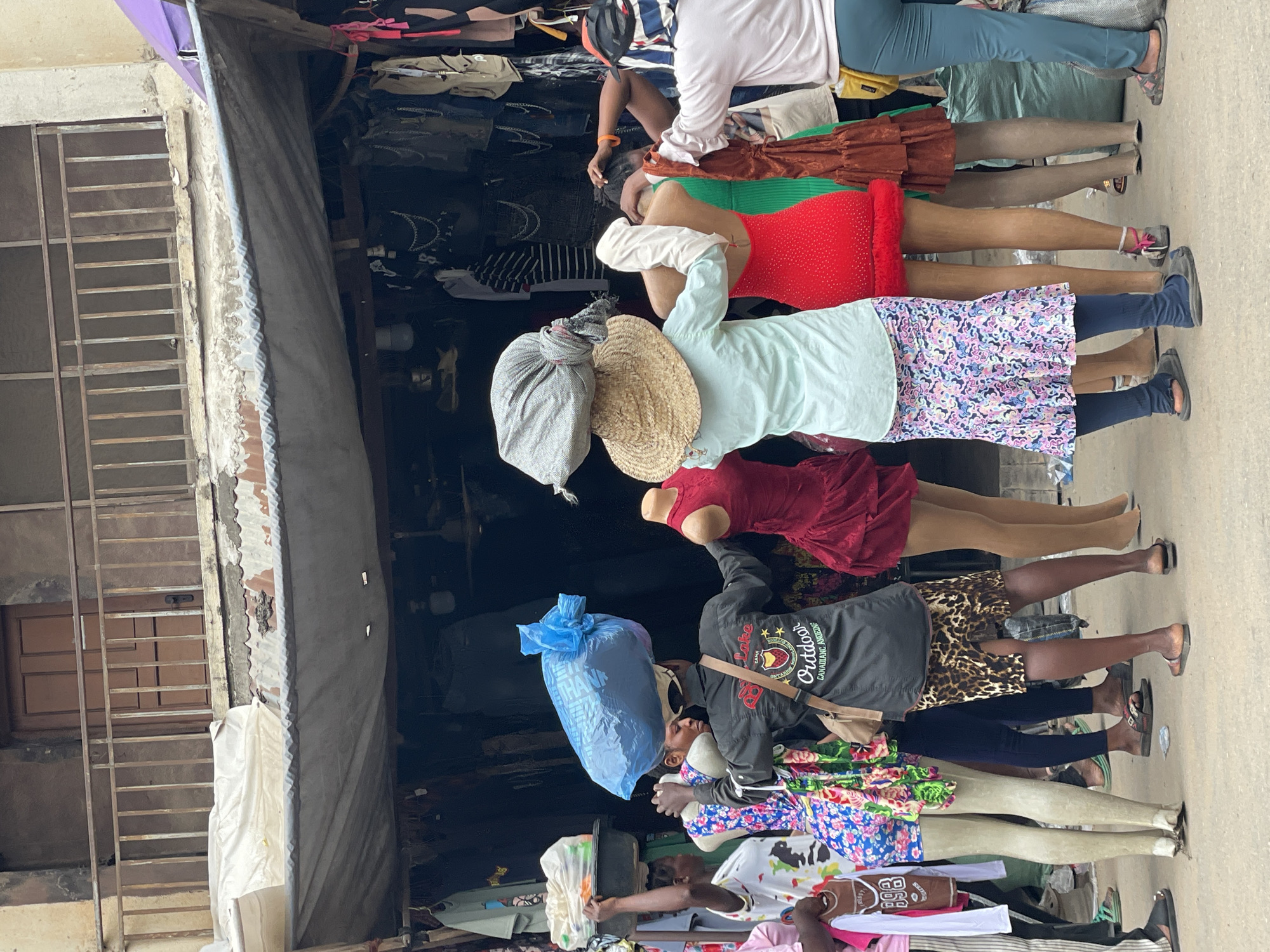 In Fegge, Nigeria, a busy outdoor market is depicted, likely selling clothing. The scene focuses on a group of people, some of whom are carrying goods on their heads. One person, viewed from behind, wears a black shirt with "Outdoor" printed on it and has a large blue plastic bag balanced on their head. Next to them, a person in a light blue shirt and a colorful patterned skirt stands with their arms around a mannequin. Several mannequins, dressed in various outfits including a red dress and a green dress, are displayed. Another person to the left carries a tray with items on their head. In the background, stalls are filled with clothing. The overall atmosphere suggests a vibrant commercial environment.