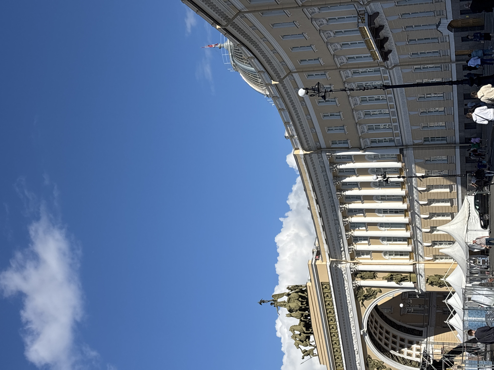 The image depicts the Palace Square in Saint Petersburg, Russia, on a clear day with scattered clouds. The iconic General Staff Building, a grand neoclassical structure with a curved facade, dominates the scene. A prominent triumphal arch, adorned with a chariot sculpture, is visible on the left side of the building.
Several people are present in the foreground, walking and standing. Some are near tents, suggesting an event or setup. A black car is parked on the right side of the square. The architecture features classical columns, ornate detailing, and numerous windows. A flagpole with a red and white flag is visible atop a dome on the building's roof. The overall atmosphere is bright and sunny, with the blue sky and white clouds providing a picturesque backdrop to the historic architecture.