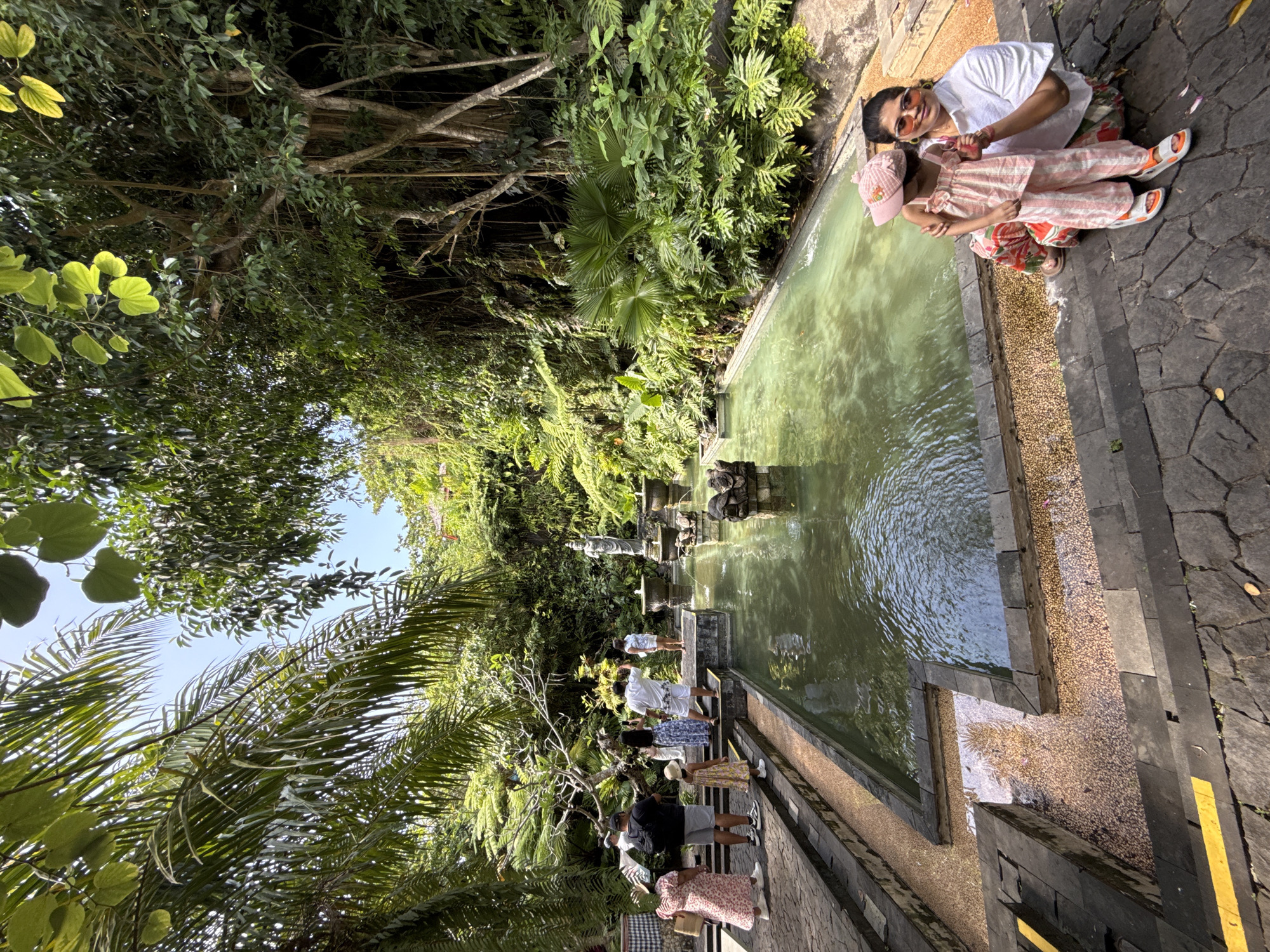 A woman and a child are photographed next to the Tirta Empul Temple holy spring water in Kuta Selatan, Indonesia. The woman wears a white t-shirt and floral-print pants, along with pink-tinted sunglasses. The child wears a pink and white striped outfit with matching hat and sandals. They are kneeling on the stone edging of the spring water pool. Several other people are visible along the length of the pool, standing near fountains of water. Lush, green foliage and trees surround the area. The photo appears to be taken at an angle, with the sky barely visible through the trees. The pool itself has green-tinted water.