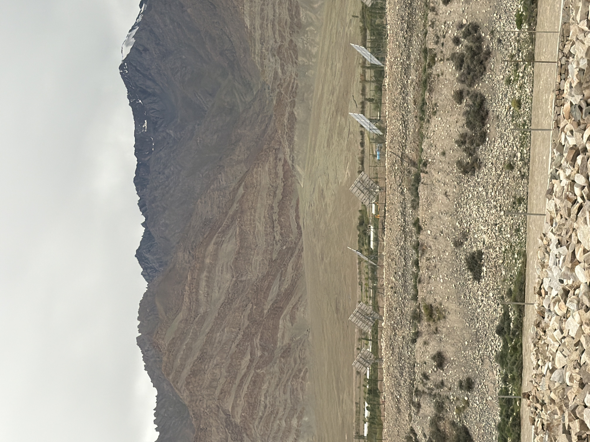 The image shows a landscape in Tanglakpurna, India, featuring a rugged mountain range in the background under a cloudy sky. The mountains display distinct geological layers of brown and tan hues, with snow-capped peaks visible on the right. In the midground, a sparsely vegetated plain stretches towards the mountains. In the foreground, a fence runs horizontally across the scene, behind which are several solar panels, some tilted towards the sky. The area in front of the fence is a rocky, barren hillside with scattered bushes. The very front of the image is dominated by a pile of large, light-colored rocks, suggesting a dry, arid environment. No people are visible in the image.
