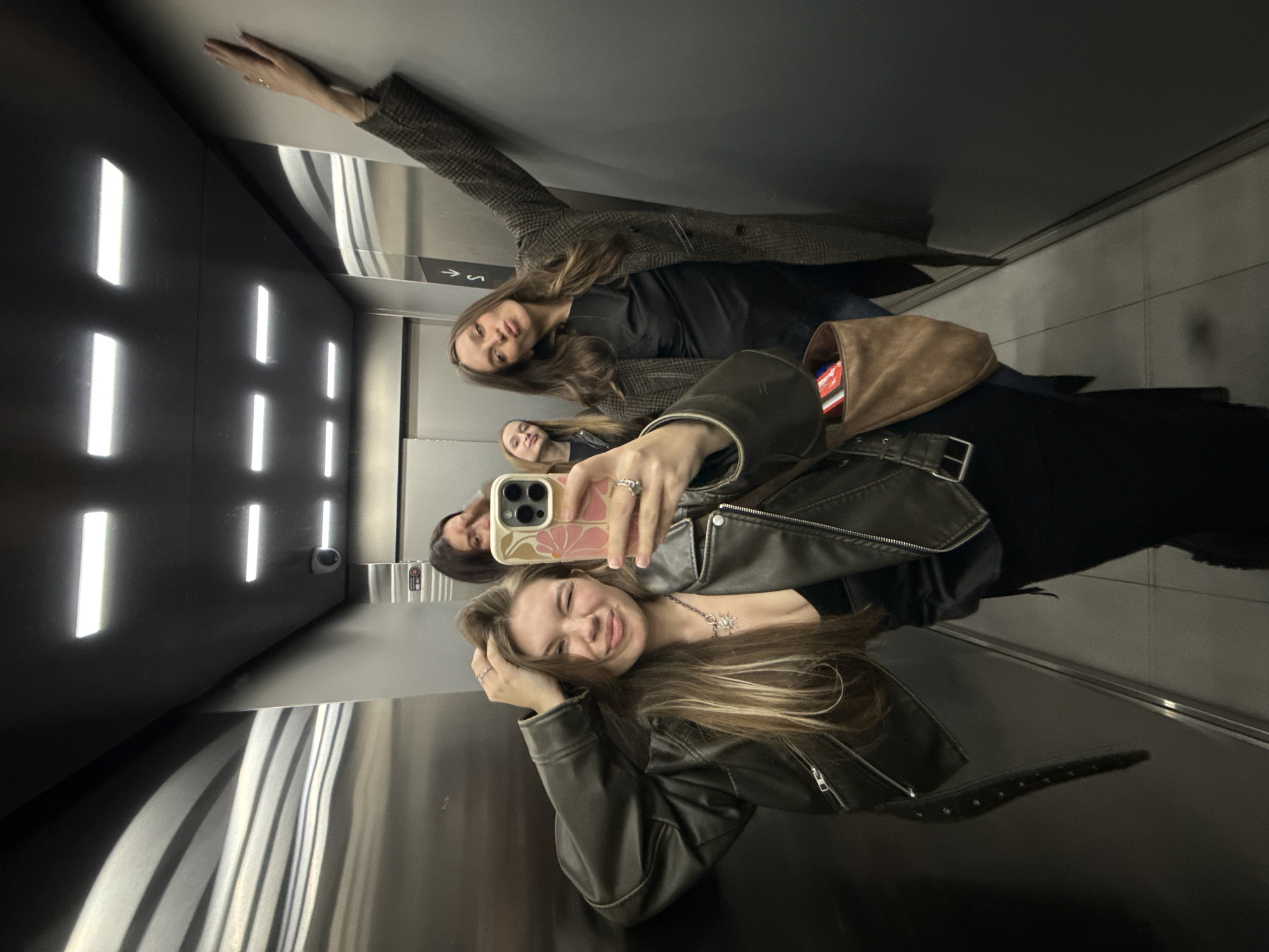 A group of four young women are taking a selfie in an elevator. The elevator has a modern design with vertical rectangular light fixtures on one wall and what appears to be brushed metal on another. The woman in the foreground is holding the phone and smiling at the camera. She is wearing a leather jacket. Behind her are three other women; one is wearing a plaid coat, and the others are partially obscured. The reflection of the women and the elevator interior indicates the photo was taken inside the elevator. The location is Belgrade, Serbia.