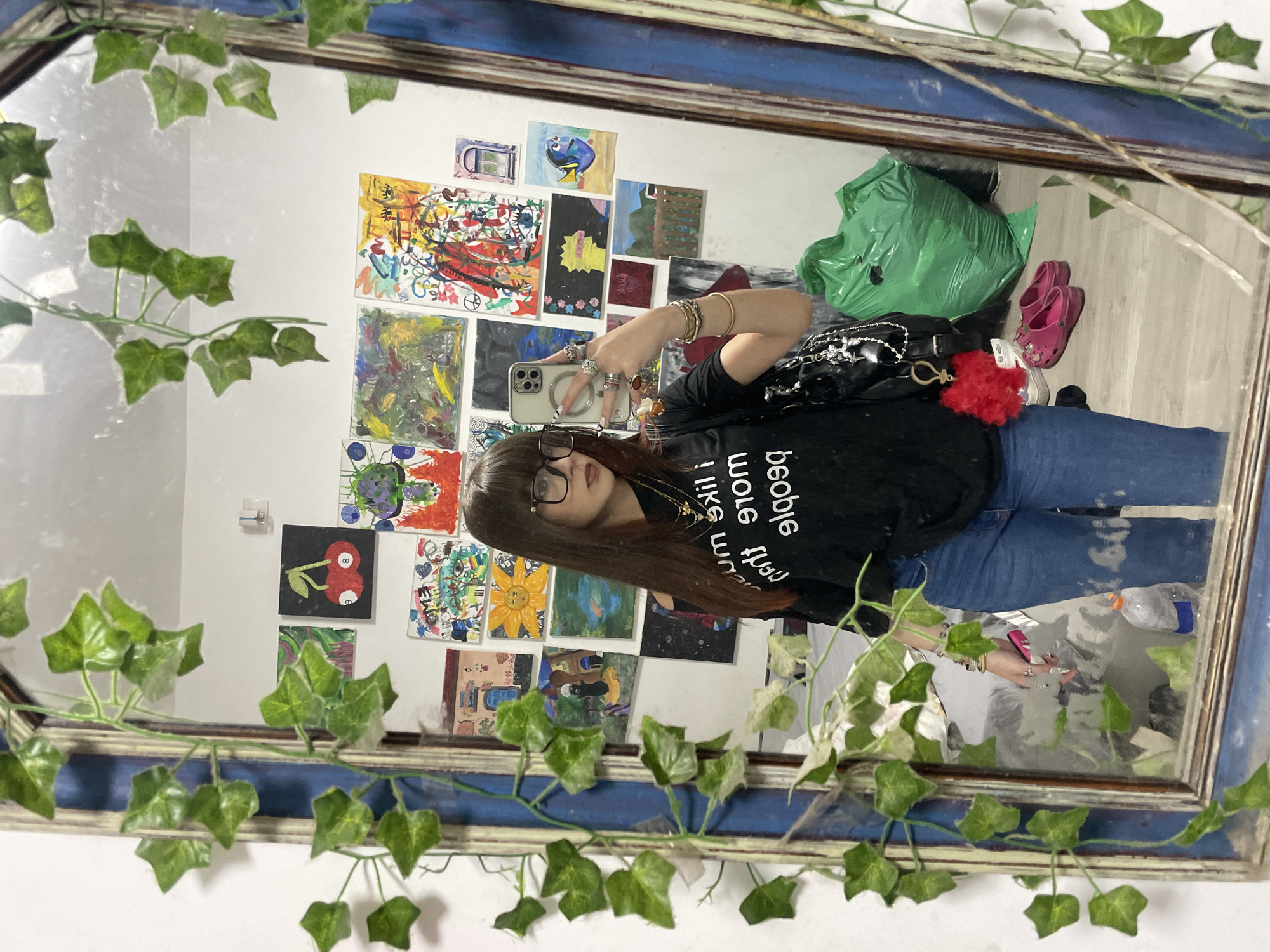 A person with long brown hair, wearing glasses, a black shirt with white lettering, and blue jeans, is reflected in a framed mirror decorated with faux ivy. The person holds a cell phone, apparently taking a photo in the mirror. Behind the person in the reflection, there is a wall covered with small paintings or drawings. On the floor in the reflection, a green bag and a pair of pink clogs are visible. A water bottle is also seen on the floor. The location is indicated as Raanana, Israel, though there are no specific features within the image to corroborate this.