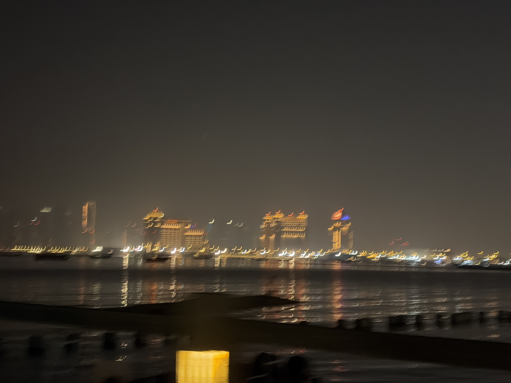 The image depicts a blurred nighttime scene of a city skyline viewed across a body of water. Several illuminated buildings are visible in the distance, appearing as streaks of light due to motion blur. The water reflects the city lights, creating a shimmering effect. The foreground shows a dark, indistinct area, possibly a waterfront or pier, with some blurred structures. There are no discernible people or specific actions taking place. The overall atmosphere is hazy and indistinct due to the low light and motion blur.