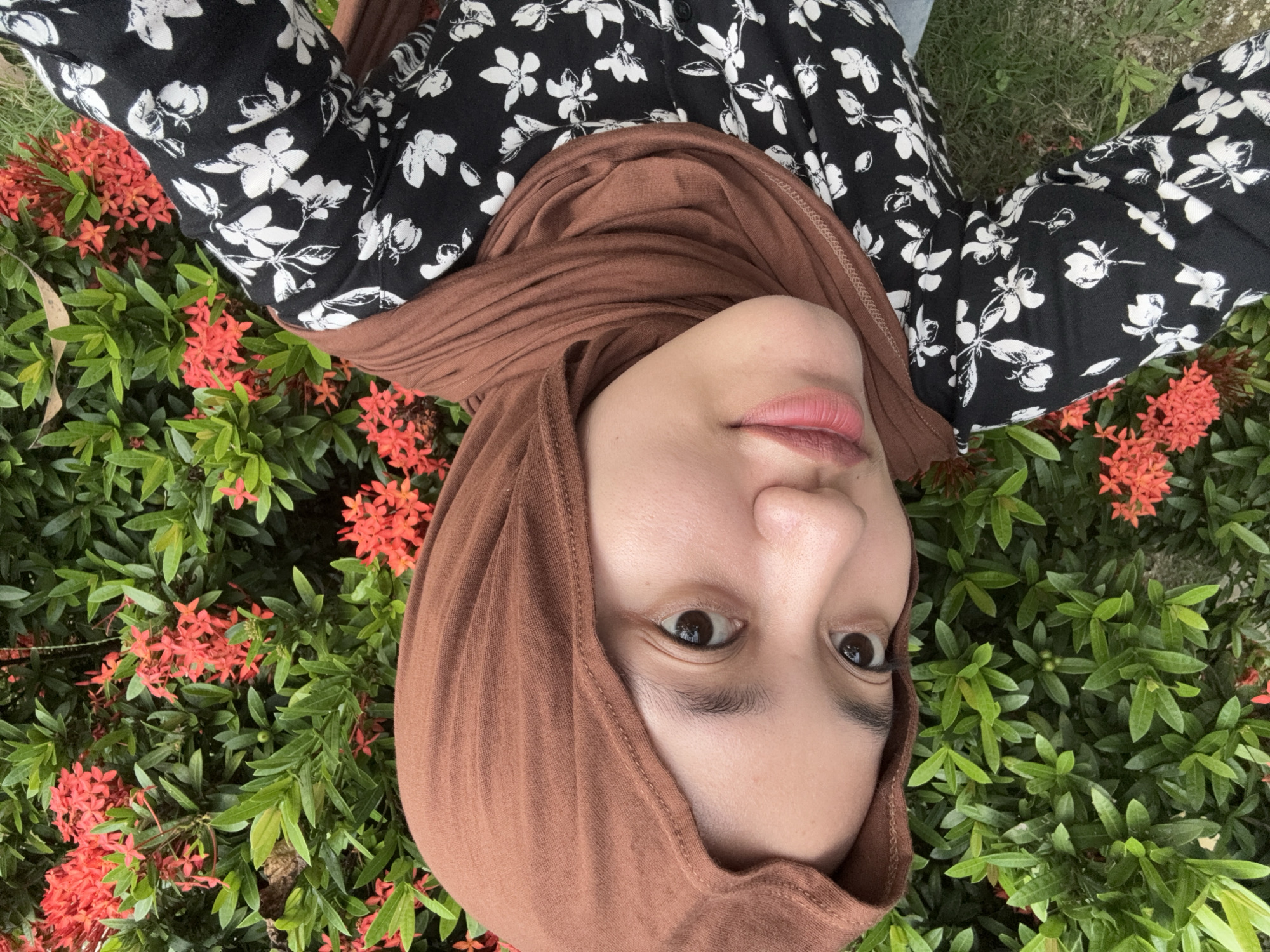 A person wearing a brown hijab and a black shirt with a white floral print is lying in a bush with red flowers and green leaves. The person's face is visible, angled towards the camera. The location is Gadingrejo, Indonesia.