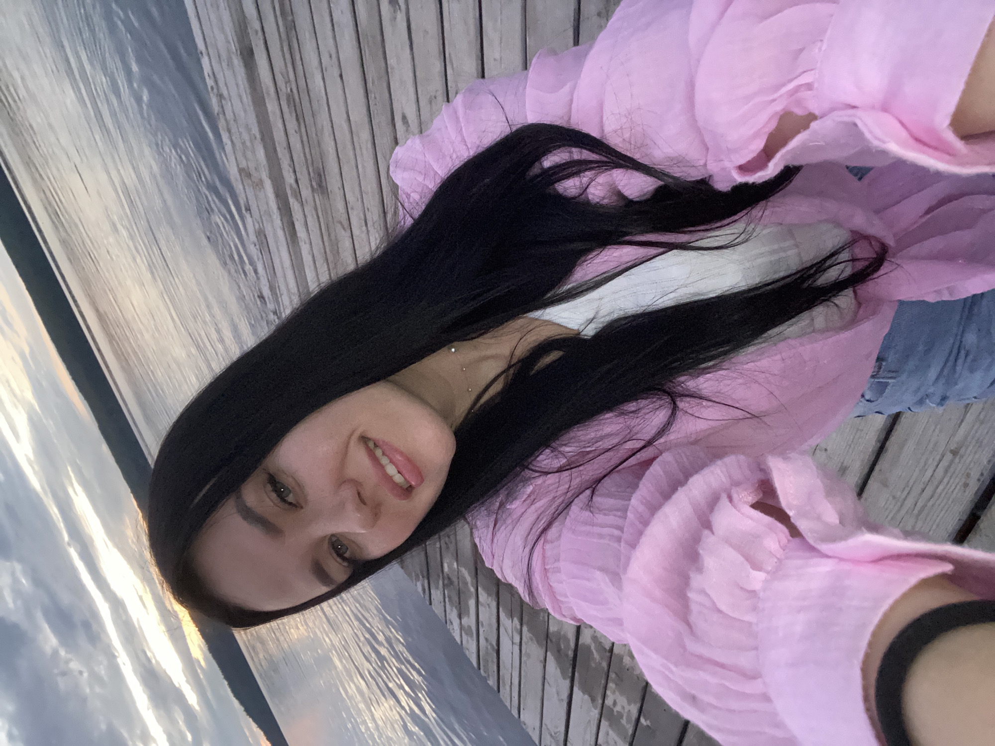 A young woman with long, dark hair smiles at the camera, taking a selfie on a wooden pier. She is wearing a light pink, sheer, ruffled blouse over a white top, and light blue jeans. The pier is made of weathered wooden planks and extends out into a body of water, likely a river or lake. In the background, the water is calm, reflecting the cloudy, twilight sky. A tree line is visible on the horizon. The location is identified as the embankment of the Kama River in Chaikovsky, Russia. The lighting suggests it is either dusk or dawn.