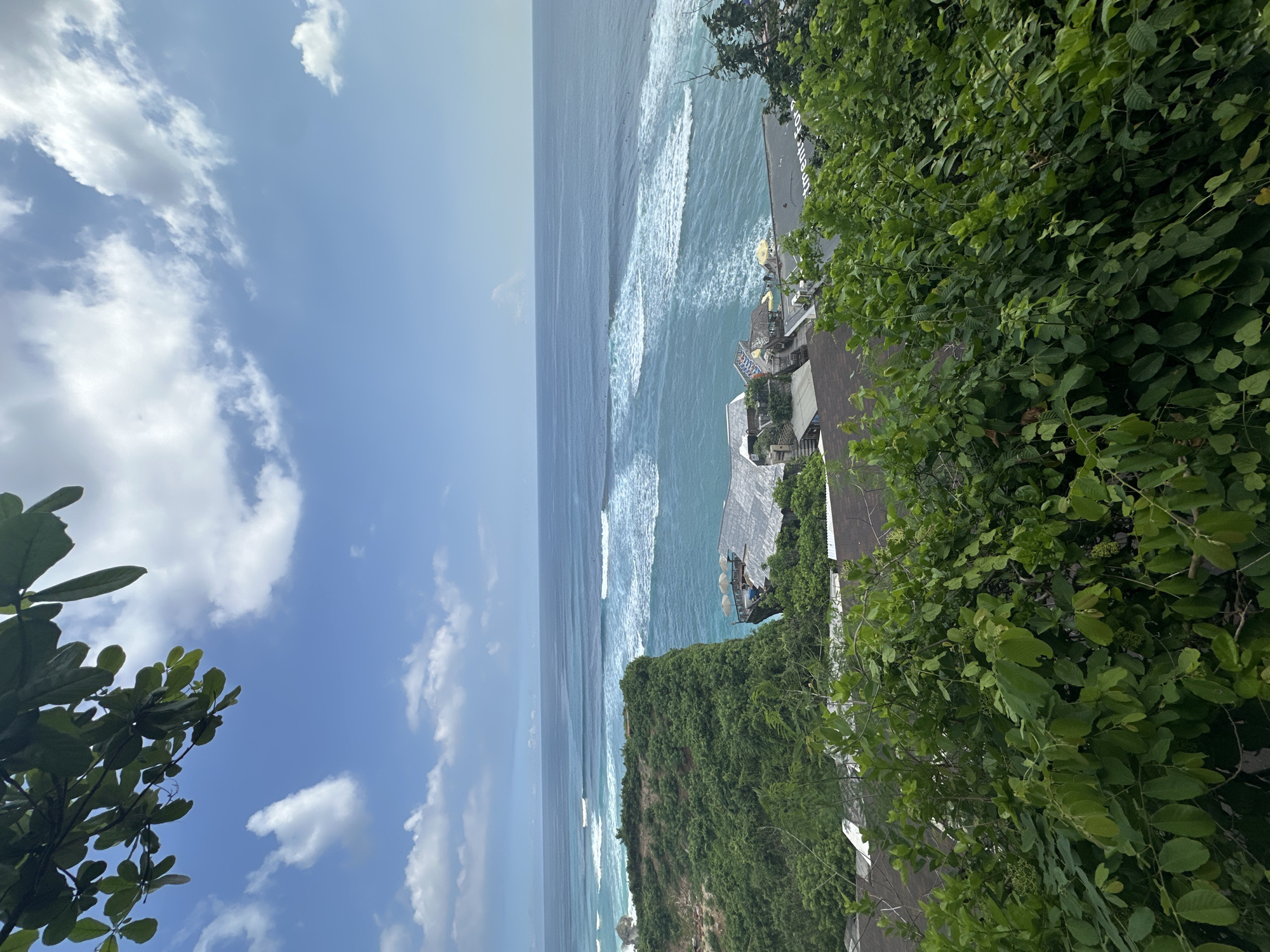 The image shows a coastal view in Kuta Selatan, Indonesia. The scene features a turquoise ocean with visible waves, a sky with scattered clouds, and a lush green cliffside covered in vegetation. On top of the cliff, there appears to be some sort of building structure with outdoor umbrellas. The image suggests a high vantage point looking down onto the ocean and the coastal landscape. The perspective is somewhat tilted.