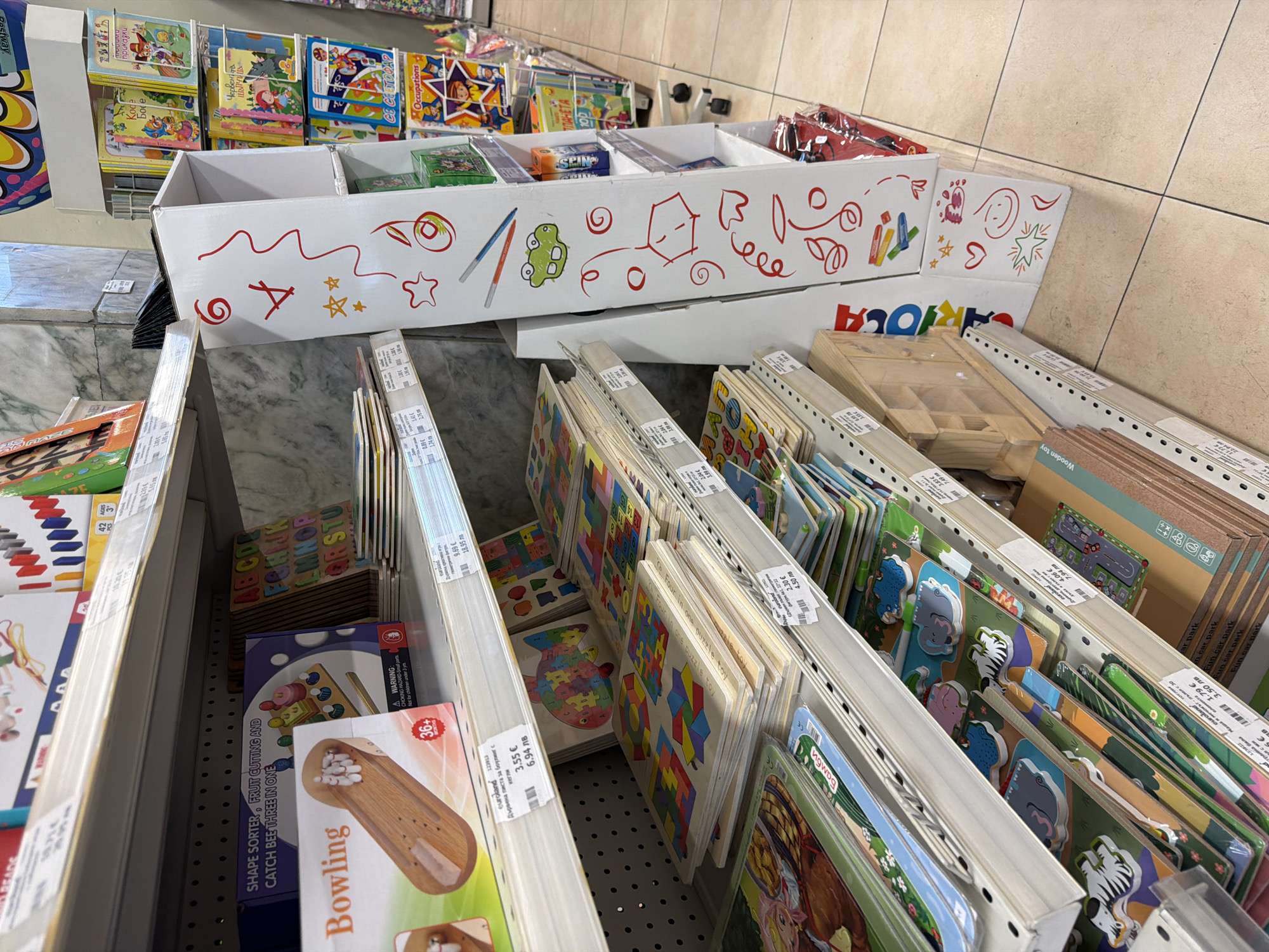 This image shows shelves of children's educational toys and books in a retail store. The store appears to be a toy store or a department store with a toy section. The items on display include jigsaw puzzles, shape sorters, and children's books with colorful illustrations. The shelves are well-stocked, and the products are neatly arranged. The overall environment is bright and clean, suggesting a well-maintained retail space. The price tags visible indicate that the items are for sale. The presence of Bulgarian text on some of the price tags and product packaging suggests that the store is located in Bulgaria. Based on the provided information, the store is located in Pazardzhik, Bulgaria. No people are visible in the image.