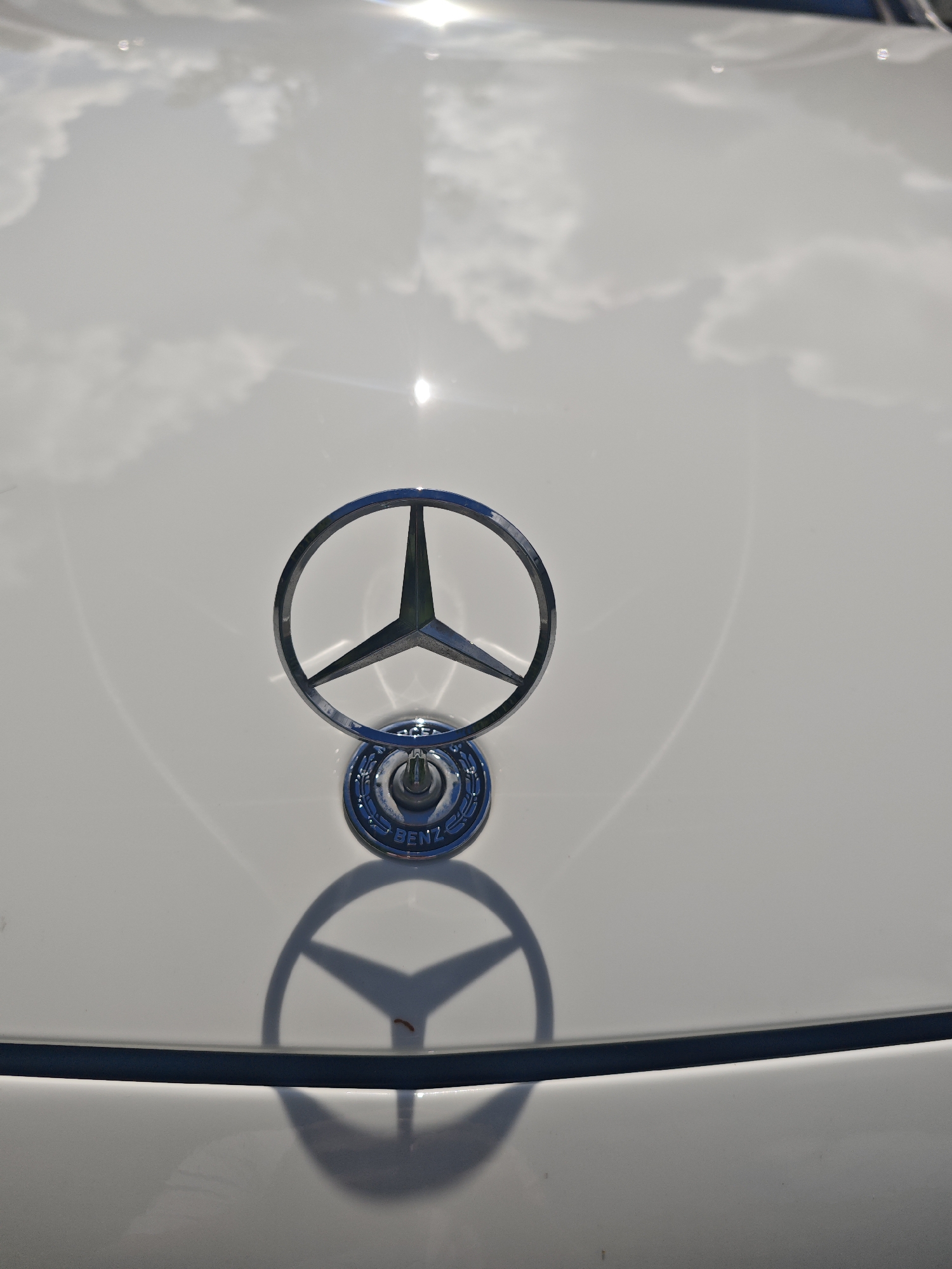 A close-up, overhead shot focuses on the Mercedes-Benz emblem on the hood of a white car. The emblem, a chrome three-pointed star enclosed in a circle, is prominently displayed. The hood's surface is highly reflective, showing a distorted reflection of a bright sky with clouds. A faint shadow of the emblem is visible on the car's hood, indicating the direction of the light source. The background is primarily the white car hood. The image does not provide any information or context related to the city of Лужники Снт, Russia, or the presence of any people or specific actions.