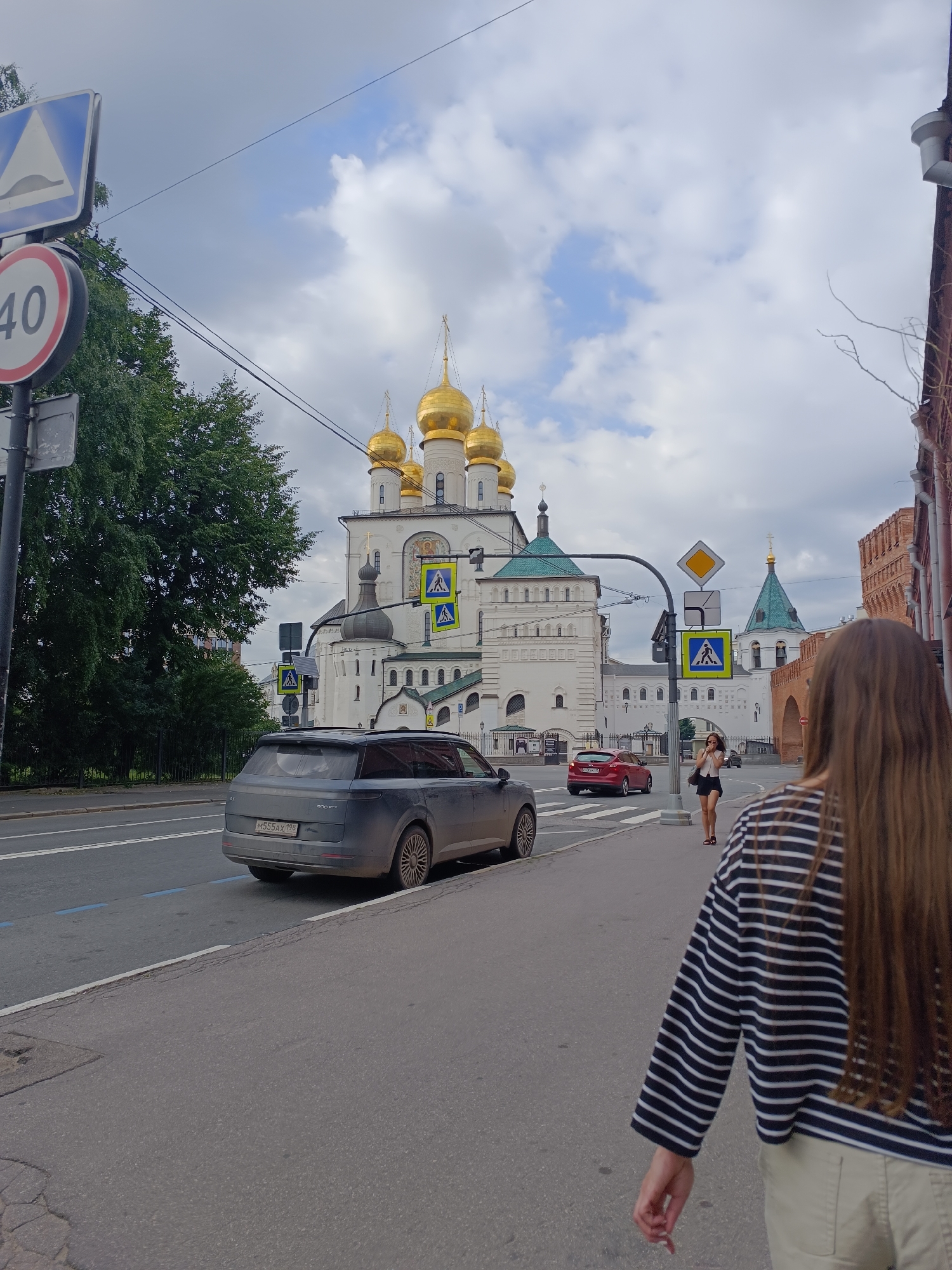 The image depicts a street scene in Saint Petersburg, Russia, with a prominent Orthodox church featuring golden domes in the background. In the foreground, a young woman with long brown hair, wearing a striped long-sleeved shirt and beige shorts, walks away from the camera along a sidewalk. To her left, a dark-colored SUV is parked by the curb. Across the street, a red car is visible, and further down the road, traffic lights and pedestrian crossing signs are present. The sky is overcast with some patches of blue sky peeking through. The architecture of the church and the surrounding buildings suggest a historical urban setting. A speed limit sign indicating 40 and a speed bump warning sign are visible on a pole to the left of the image.