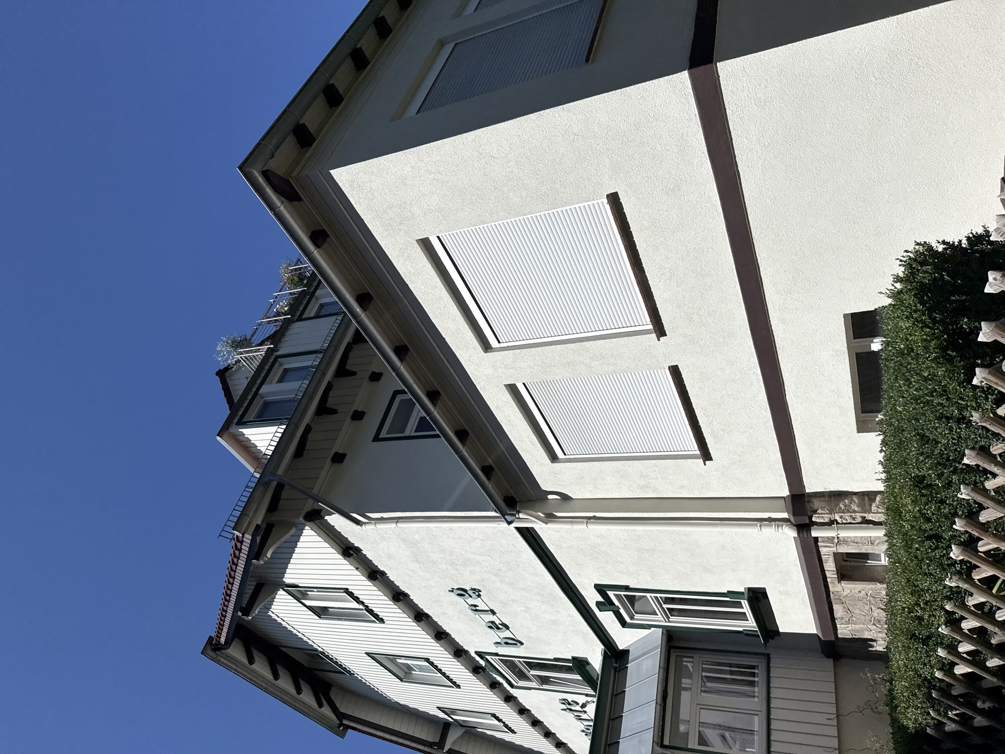 The image shows a three-story building with white stucco siding and dark brown trim. The building has a pitched roof with red tiles and white wooden eaves. There are several windows on the facade, some with green trim and others with white roll-up shutters. A sign with green lettering is visible on the building, partially obscured by shadow, but "berg" is clearly legible. The building appears to be a residential or commercial property. In the foreground, there is a neatly trimmed green hedge and a rustic wooden fence. The sky above is a clear, bright blue. The image appears to be taken on a sunny day. The building is identified as part of the "Haus am Berg" in Bad Sachsa, Germany, a guesthouse with a restaurant.