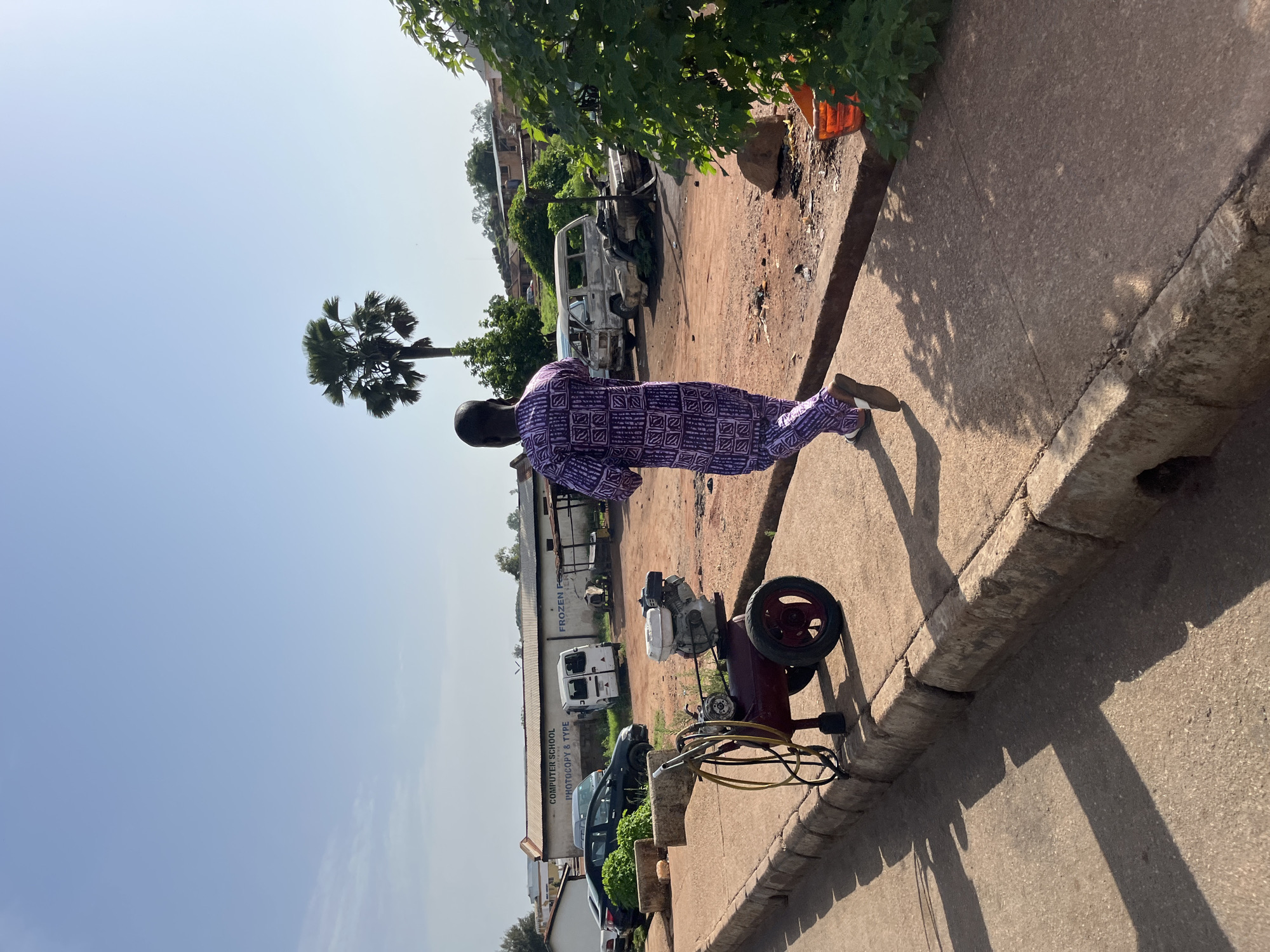 A person, dressed in a purple and white patterned robe, stands on a sidewalk next to a dusty road. The person's back is to the camera, and they appear to be looking at a red and black air compressor with a yellow hose, which is positioned on the sidewalk. Behind the person, there's a building with signage that reads "COMPUTER SCHOOL" and "PHOTOCOPY & TYPE" and "FROZEN FOOD". A white van and other vehicles are parked in the background, along with some trees and buildings. A palm tree is visible to the left of the frame. The scene appears to be outdoors during the day, with a clear blue sky and shadows cast by the sun, suggesting it is a sunny day. The environment seems to be a commercial area or a street in a town.