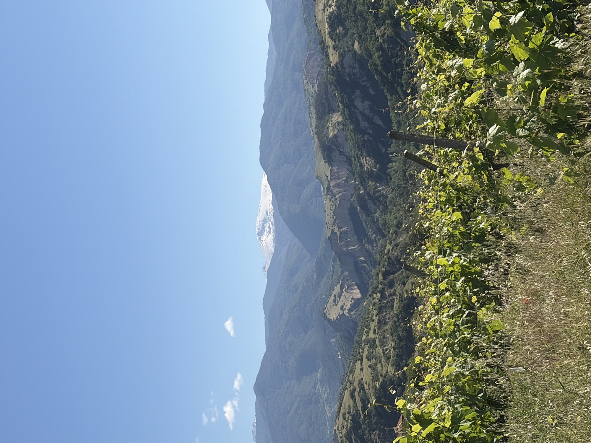 The image depicts a mountainous landscape under a clear blue sky. In the foreground, there is a lush area of greenery that appears to be vegetation. The mountain range spans the midground, with peaks reaching into the sky, partially covered with snow. There is a cloud or clouds present in the sky. The location is Unknown, Unknown.