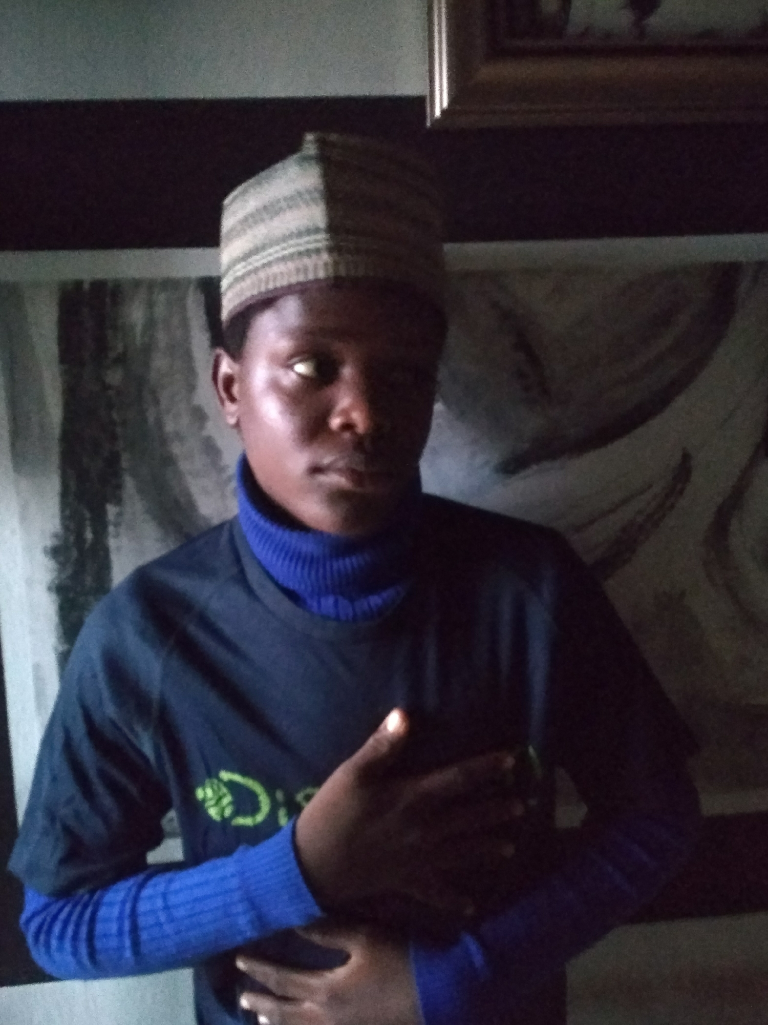 A person stands indoors. They are wearing a light brown cap, a dark blue t-shirt over a bright blue turtleneck sweater. The person has dark skin. Their hands are clasped in front of them. Behind the person is a partially visible wall decoration. Above the decoration is a dark horizontal band. The scene is dimly lit. There is no verifiable evidence from the image that indicates this scene occurred in Jos, Nigeria.