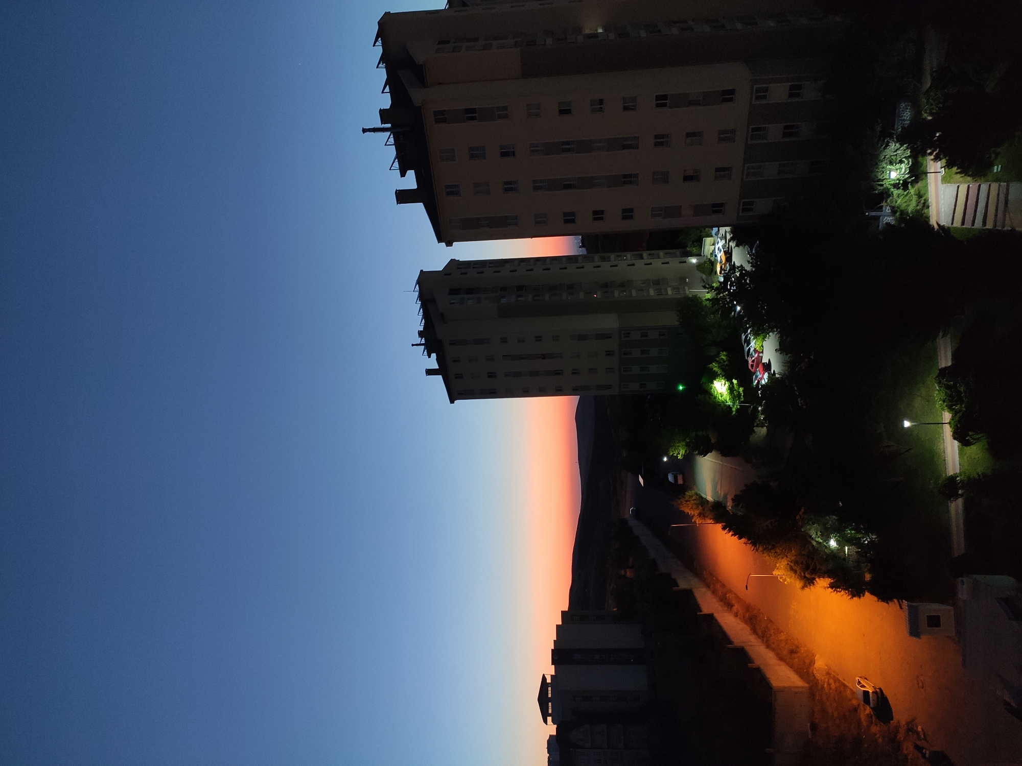 The image captures a twilight scene in Etimesgut, Turkey, featuring two tall apartment buildings as the focal point. The sky transitions from a deep blue at the top to hues of orange and pink near the horizon, indicative of sunset or dawn. In the foreground and midground, the apartment buildings are silhouetted against the fading light. To the left, other lower buildings and a street are visible, illuminated by streetlights and the warm glow from a street lamp. Several cars are parked along the street and in a lot adjacent to one of the buildings. Trees and other foliage dot the landscape, some catching the artificial light. The overall impression is a residential urban environment transitioning from day to night.