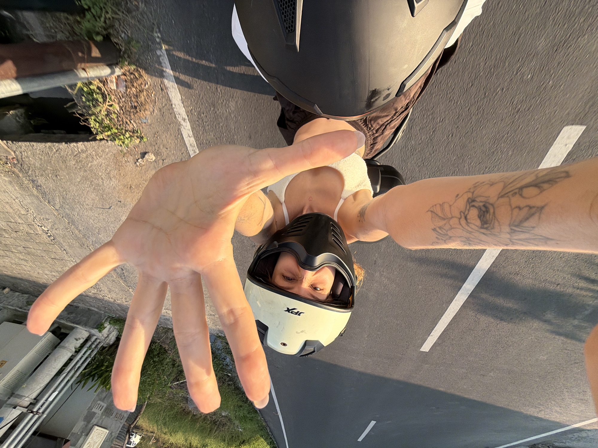 A person is riding a motorcycle on a paved road in Mengwi, Indonesia, and taking a selfie. The person is wearing a white and black helmet and a white top. Their left arm, with a tattoo of flowers, is visible. The right hand is held up towards the camera, obscuring part of the face. The road has white markings and is bordered by grass and a ditch on the right. A white car is visible in the background on the left side of the road. Electrical conduits and a building facade are also visible on the right side of the road. The lighting suggests it is late afternoon or early evening.