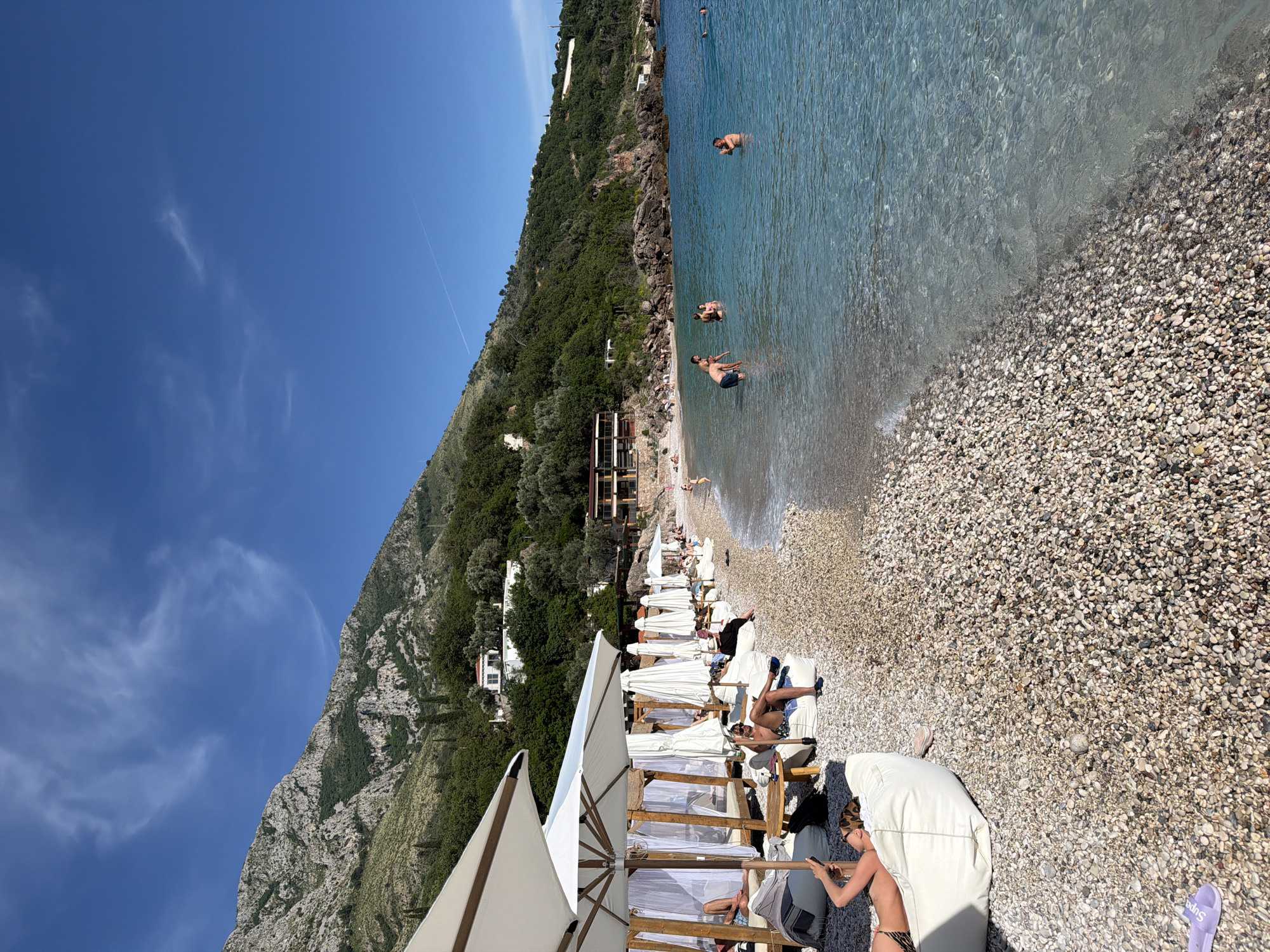 The image shows a beach in Sveti Stefan, Montenegro. The beach is made of small pebbles and has clear, turquoise water. There are people swimming and sunbathing in the water. On the beach, there are rows of white lounge chairs and umbrellas. People are relaxing on the chairs. Behind the beach is a steep, green mountain with some buildings on its slopes. The sky is blue with a few wispy clouds. A contrail is visible in the sky.