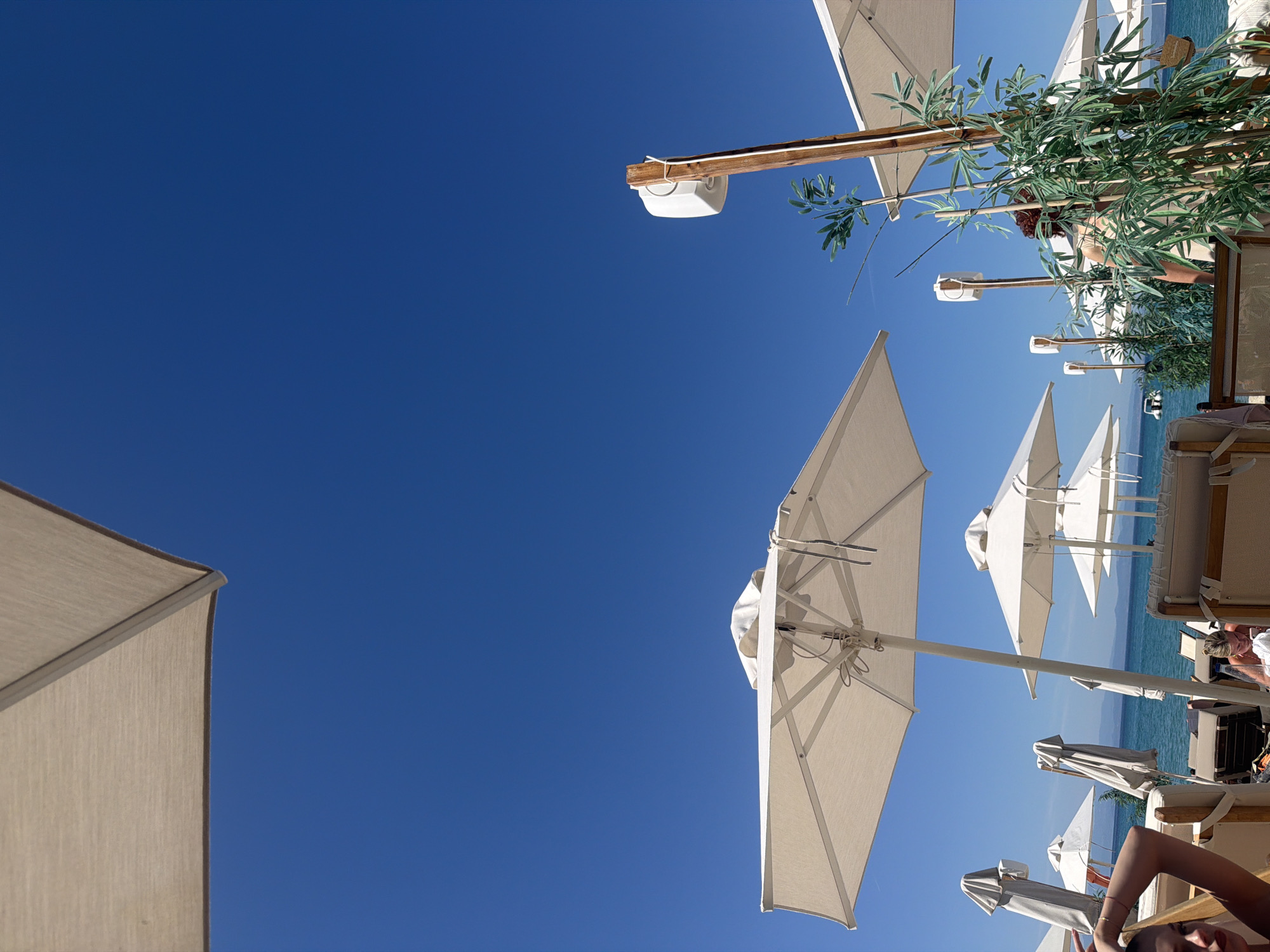 The image shows a beach scene in the city of Pallini Municipal Unit, Greece. Several white beach umbrellas are visible against a clear blue sky. Some beach chairs are present, with at least two people lounging on them. The sea is visible in the background. There are some green plants near some of the umbrellas.