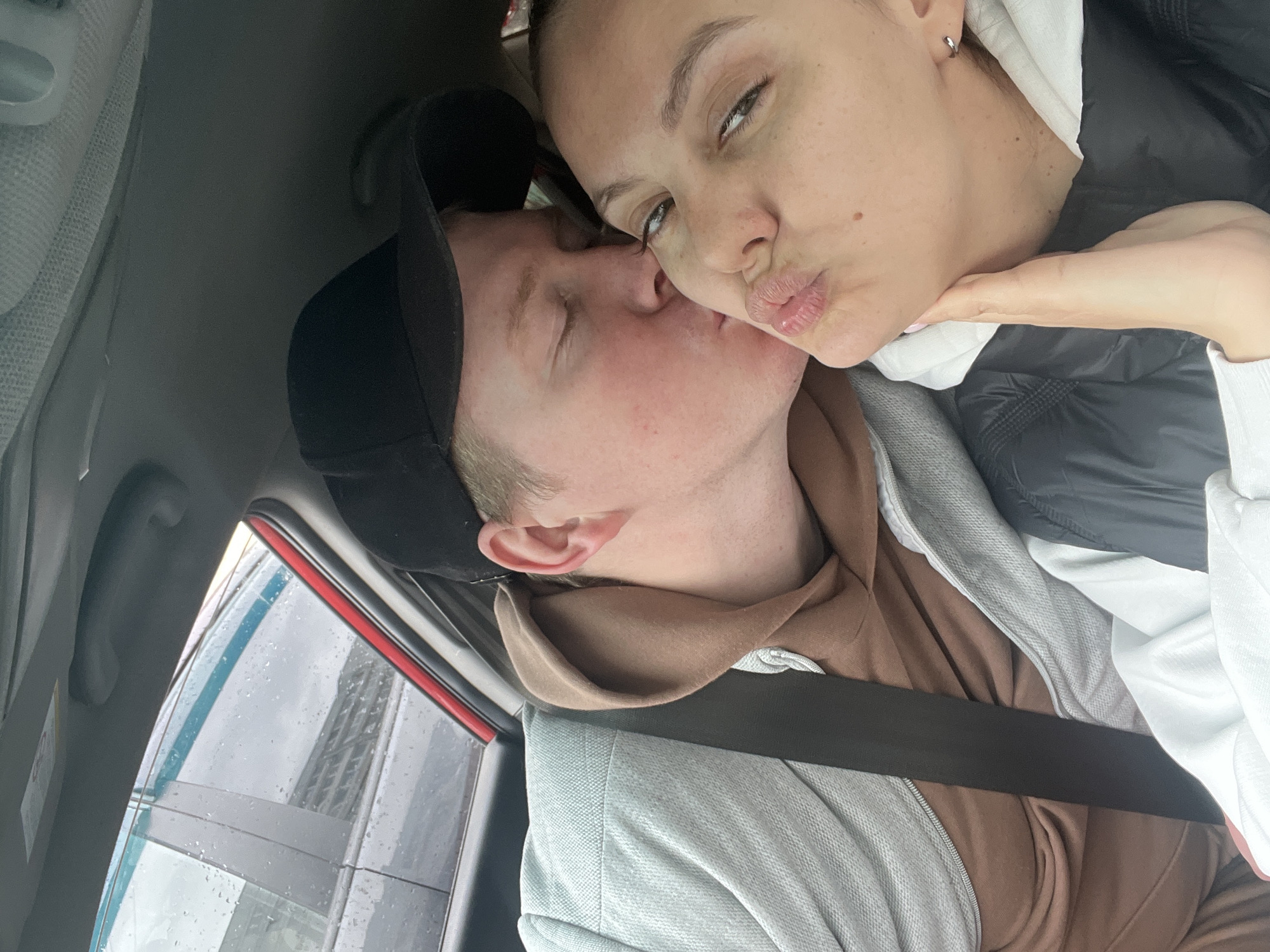 Inside a car, two individuals, a male and a female, are sharing a tender kiss. The image is taken at a slight angle, giving an overhead perspective. The male, positioned on the left, wears a black baseball cap, a light brown hooded sweatshirt, and a light grey polo-style shirt. His eyes are closed as he leans in to kiss the female on the mouth or cheek. The female, on the right, wears a black and white hooded jacket, with a white garment visible underneath. She has an arm resting against her chin, and is making a pouting face for the kiss, with one eye partially open, looking towards the camera. She has a small silver hoop earring in her visible ear.
Raindrops are visible on the car's side window, suggesting rainy or overcast weather. Through the window, a large, tall structure, possibly a building or tower with a distinct grid pattern, is partially visible in the background. A dark seatbelt is seen across the male's chest. The car's interior, including parts of the headliner and window trim, is also visible. The scene exudes a sense of intimacy and affection between the couple, captured in Chaikovsky, Russia. There is no discernible text visible in the image.