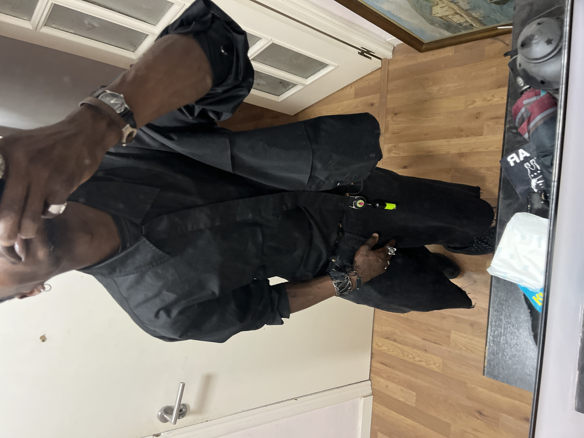 The image captures a person indoors, dressed in an all-black outfit and adorned with striking accessories, likely taking a photo. The individual has a dark skin tone, with their head partially visible on the left, looking downwards. They wear a silver-toned watch with a light band on their left wrist, multiple rings on both hands (several prominent on the left, one on the right pinky), and several dark and silver-toned bracelets on their right wrist. A small, bright green object with a black cap and clip is attached to their clothing near their waist.
The person stands on a light-colored wooden floor. Behind them on the left is a plain light-colored wall with a silver-toned door handle visible at the bottom. In the upper background, a white door with two frosted glass panes is slightly ajar. Above this door, a framed painting depicting a ship or large vessel on water is partially visible.
To the right of the person, a dark counter or shelf holds several items, including a white plastic bag, a dark metallic teapot-like object, and a crumpled red and black striped fabric. A black item with reversed white text, "RA" and possibly part of "BBIT", is also on the counter, suggesting the image might be a mirror reflection from the far right edge. The setting appears to be an interior space, such as a home or entryway, with ambient indoor lighting. The location context provided is Barcelona, Spain.