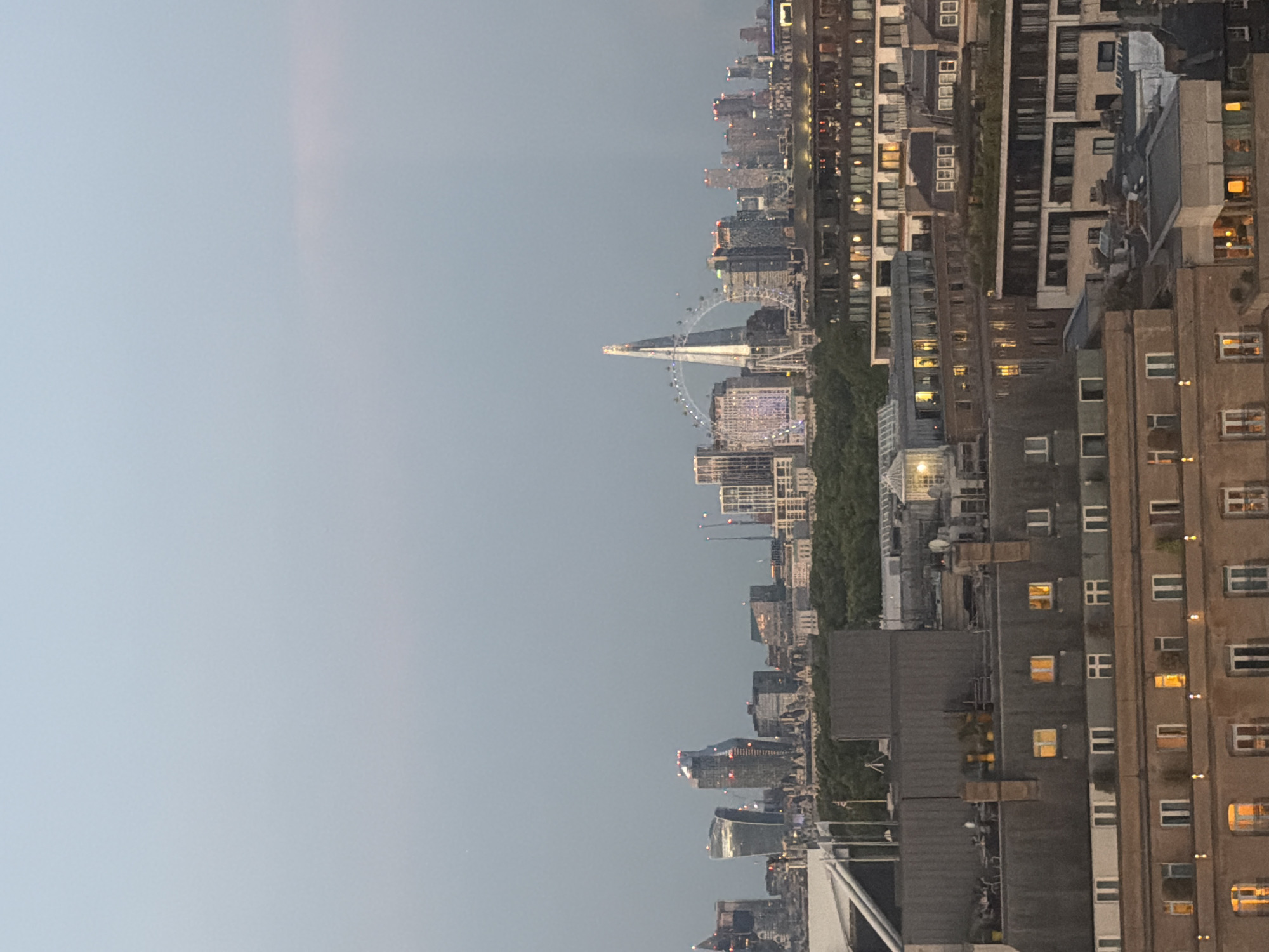 The image displays a cityscape of London, United Kingdom, from a high vantage point, likely at dusk or early evening. The sky is a pale, overcast blue. In the middle ground, a cluster of modern skyscrapers dominates the skyline, including the distinctive shard-shaped building and the London Eye Ferris wheel. The foreground is filled with the roofs and upper floors of several buildings, many with illuminated windows, suggesting residential or commercial spaces. Trees are visible between the foreground buildings and the distant skyscrapers, forming a green band across the middle of the image. The overall atmosphere is one of urban development and activity at the close of the day.