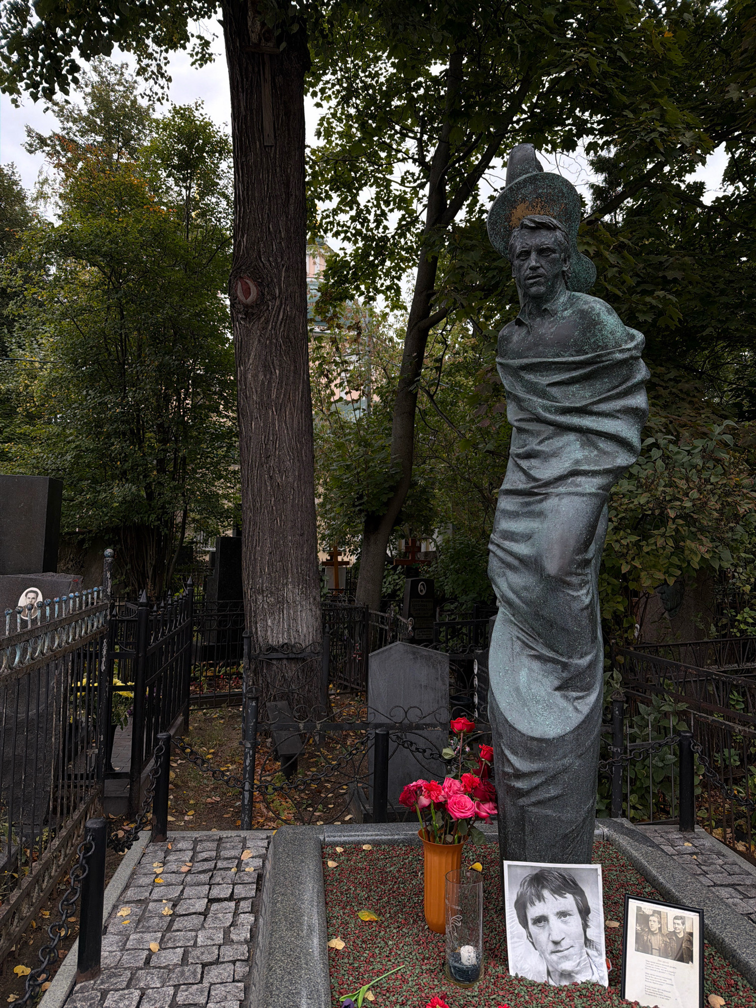 The image depicts a gravestone at Novodevichy Cemetery in Moscow, Russia. The gravestone features a bronze statue of a man wearing a hat and draped in fabric, standing next to a tall tree. In front of the statue are a vase of red roses, a tall glass with a candle, a framed photograph of a man, and another framed photograph of two young men. The ground is covered with red gravel and scattered autumn leaves. The cemetery is surrounded by black metal fences and other gravestones and trees are visible in the background.