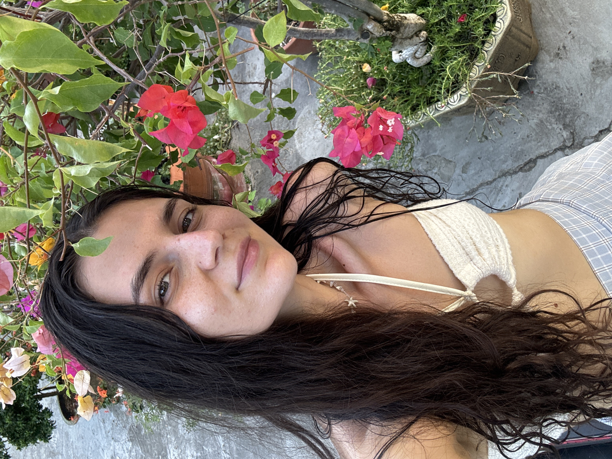 A young woman with long, dark hair and a fair complexion is positioned at an angle in the frame, surrounded by greenery and flowers. She is wearing a white, knitted top with thin straps and what appears to be plaid pants. She appears to be in an outdoor setting with potted plants including bougainvillea. The setting seems to be a patio or garden area with concrete flooring. The location is Mũi Né, Vietnam.