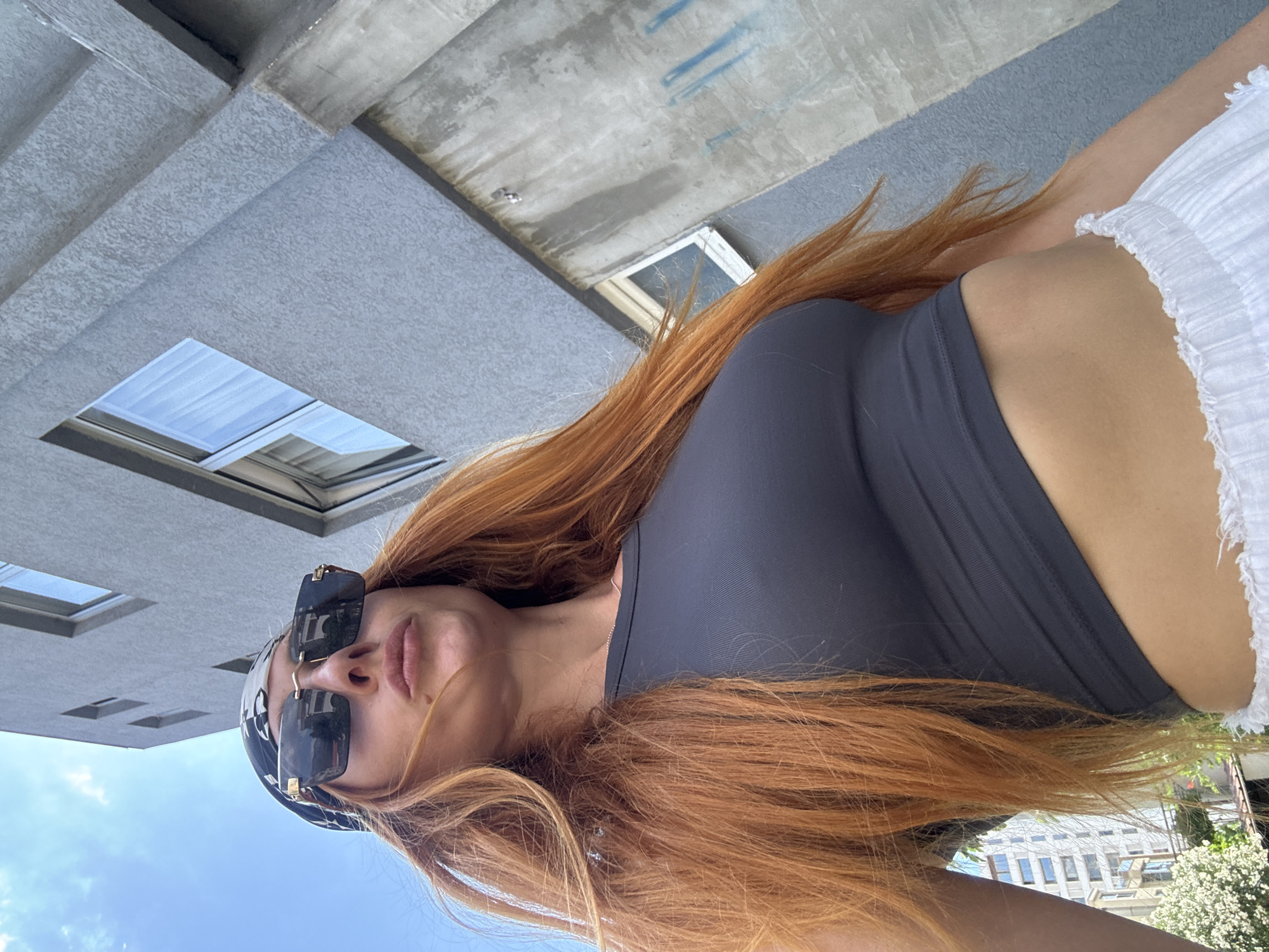 A low-angle, full-body portrait shows a woman with long, auburn hair posing outdoors. She is wearing a dark grey tank top and white, ruffled shorts. Large, dark-lensed sunglasses with gold accents are perched on her nose, and she has a bandana on her head. The background features a grey building with visible windows and concrete elements, suggesting an urban or architectural setting. The sky above is a clear blue, indicating it's daytime with good weather. The image is oriented vertically, with the woman's body stretching from the top to the bottom. The overall mood appears casual and sunny, possibly taken on a vacation or during a warm-weather outing.