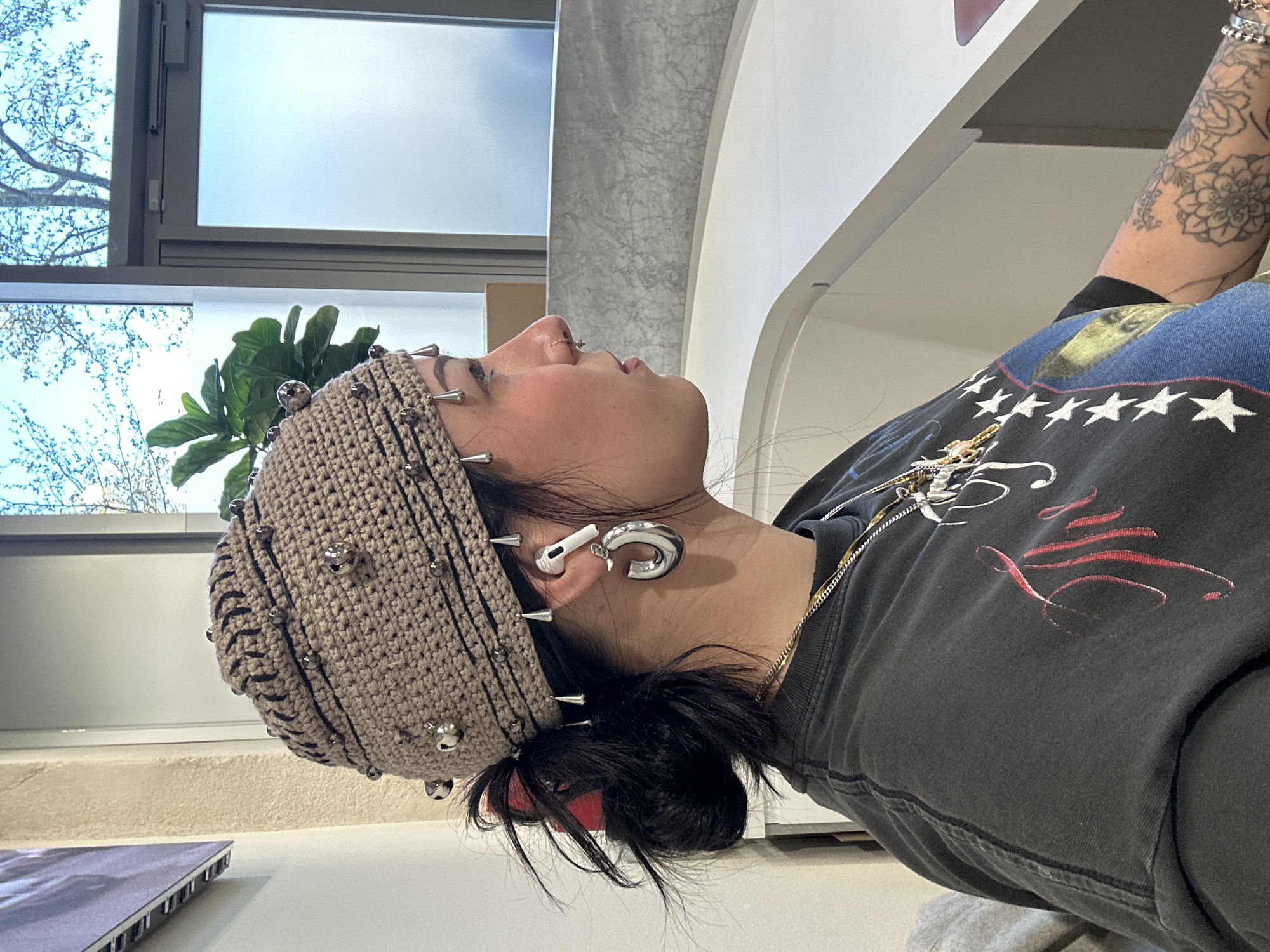 A woman with a nose ring and multiple ear piercings wears a crocheted hat with studs or spikes, an AirPod in one ear, and a large silver earring in the other. She is wearing a black t-shirt with a design featuring stars and writing. Part of a tattoo is visible on her arm. The setting appears to be indoors, possibly a store or office in Paris, France, judging from the architectural style of the window frames and interior design elements. The lighting is bright and natural, suggesting it is daytime. A plant is visible near the window.