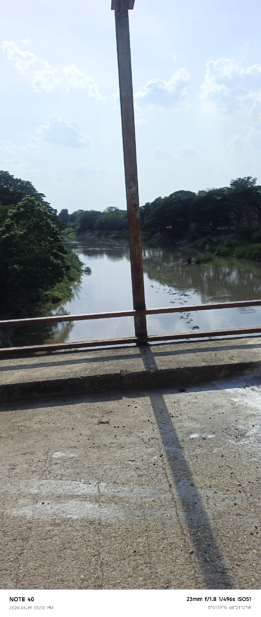 Here is a description of the image:
The image captures a view from a bridge over a muddy river in El Baúl, Venezuela. The bridge's concrete surface takes up a large portion of the foreground. A rusty metal post stands prominently, casting a dark shadow onto the bridge deck. The river is flanked by dense vegetation along its banks, with trees visible on both sides. The sky above is a mix of blue with scattered clouds. The photo was taken on April 19, 2026, at 3:50 PM. The camera settings were 23mm focal length, f/1.8 aperture, 1/496 second shutter speed, and ISO 51. The GPS coordinates are 8°41'59"N 68°21'12"W.