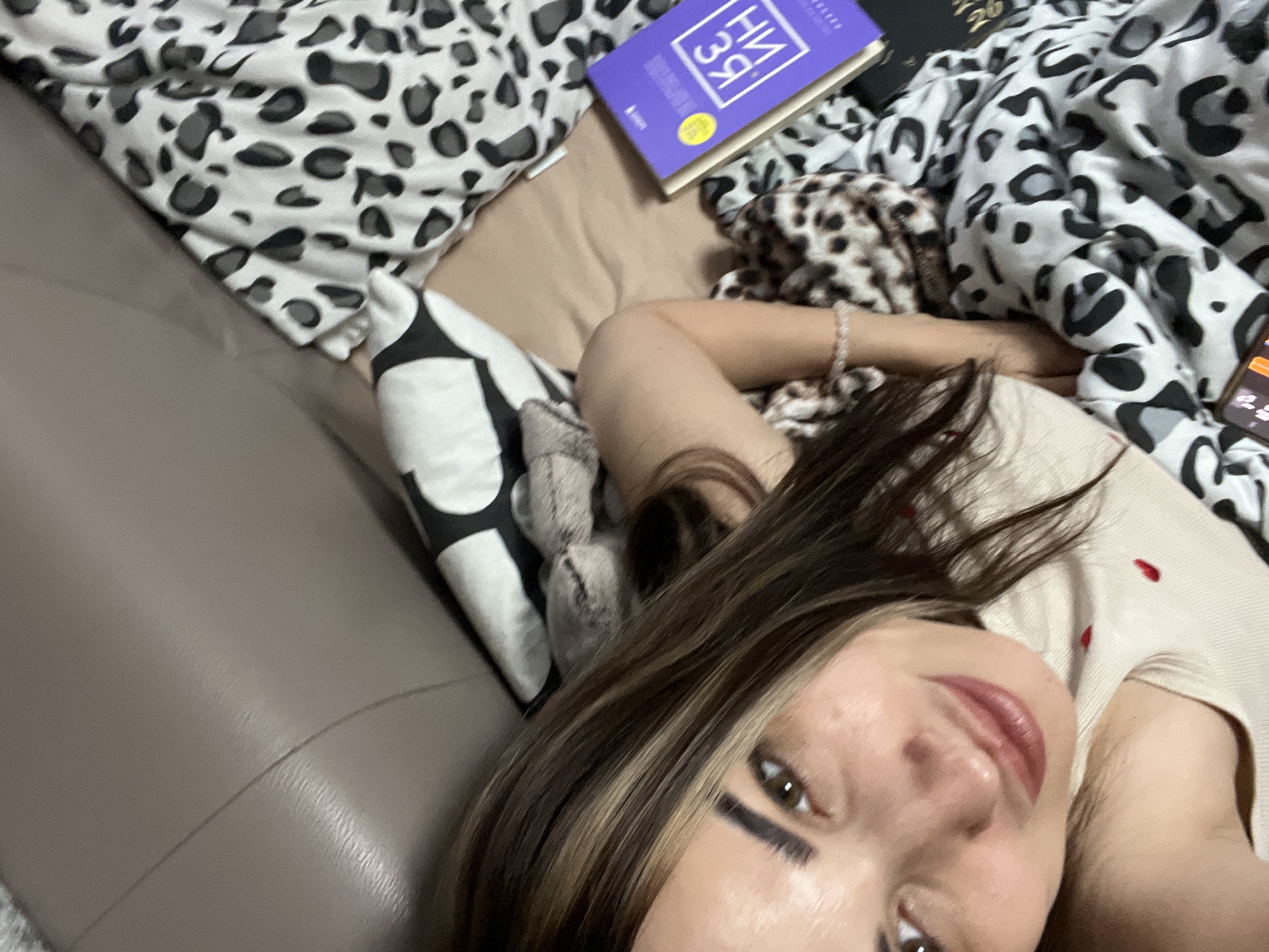 A young woman is depicted in a close-up, upward-angled shot, lying in a relaxed pose and looking directly at the camera. She has long brown hair with prominent lighter highlights, styled to fall to her left. Her makeup includes thick black winged eyeliner and reddish-brown lipstick. She is wearing a light-colored top, possibly beige or off-white, adorned with small red heart-like details. A light-colored beaded bracelet is visible on her right wrist.
She is positioned on what appears to be a couch or bed, partially covered by a prominent black and white leopard print blanket or duvet. Another smaller item with a brown and black leopard print is near her right arm. To her left, a black and white patterned pillow is visible. The background includes a light brown upholstered surface, likely the arm or backrest of a couch.
Near her left arm, a purple-covered book lies face-up on the blanket. The cover displays text in Cyrillic script, with "НИ ЗЯ" clearly visible, along with a yellow circular sticker. A dark-covered book is partially seen beneath or adjacent to the purple one. A smartphone with its screen visible rests on the leopard print blanket near her right arm. The overall scene suggests a moment of leisure and comfort indoors.