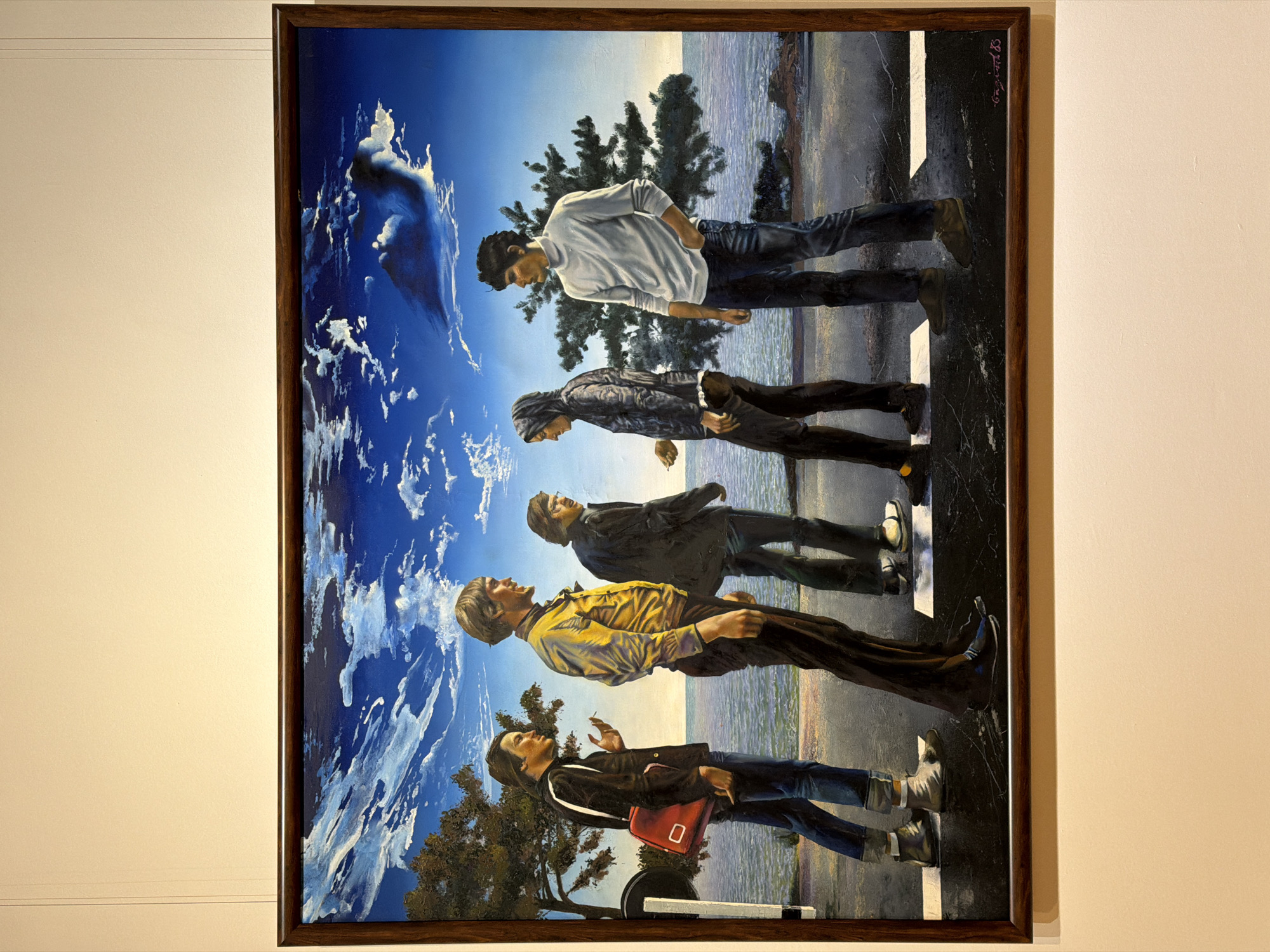 The image depicts a framed oil painting, hung on a light beige wall. The painting shows four men walking in single file across a crosswalk in what appears to be Moscow, Russia. The background transitions from a blue sky and cloudscape at the bottom to a body of water at the top. Each man is wearing different clothing: the man at the bottom wears a black jacket, blue jeans, white sneakers, and carries a red satchel. The next man wears a yellow shirt, dark pants, and dark shoes. The third man wears a black jacket, blue jeans, and dark shoes. The top man wears a white sweatshirt, blue jeans, and dark shoes. The crosswalk is white and contrasts against the dark asphalt. A tree appears to be located in the lower-left corner. The painting is signed in the top-right corner.