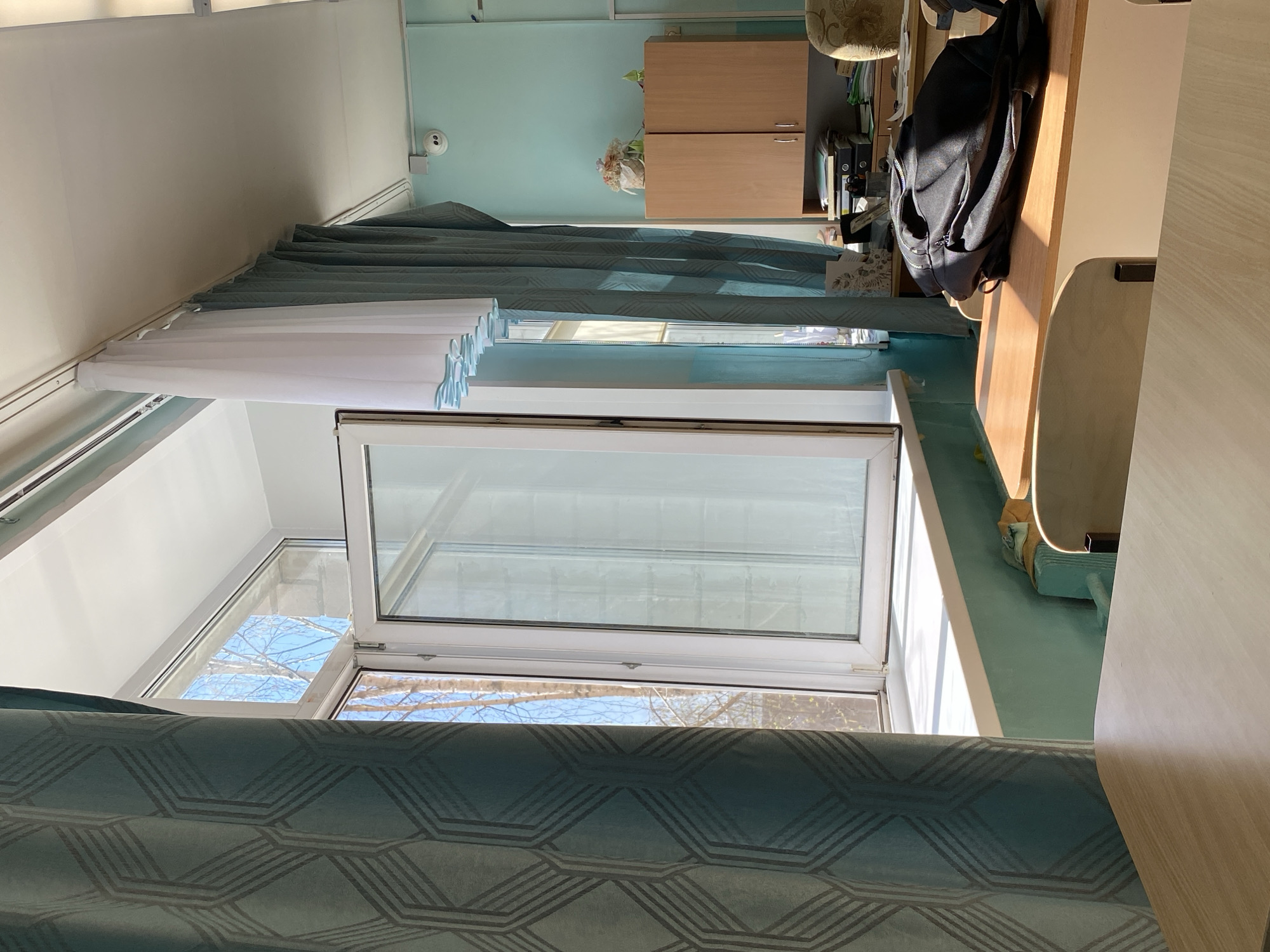 The image shows an interior view, likely of a room, with a window partially open and curtains drawn. The location is presumed to be Votkinsk, Russia, based on given information, although this cannot be verified from the image itself. The window has a white frame, and the view through the open portion shows trees against a sky with some clouds. The curtains are blue with a geometric pattern. A portion of what appears to be a cabinet is visible in the background, along with a desk and a backpack. The room's walls are painted in a light blue hue. No people are visible in the image.