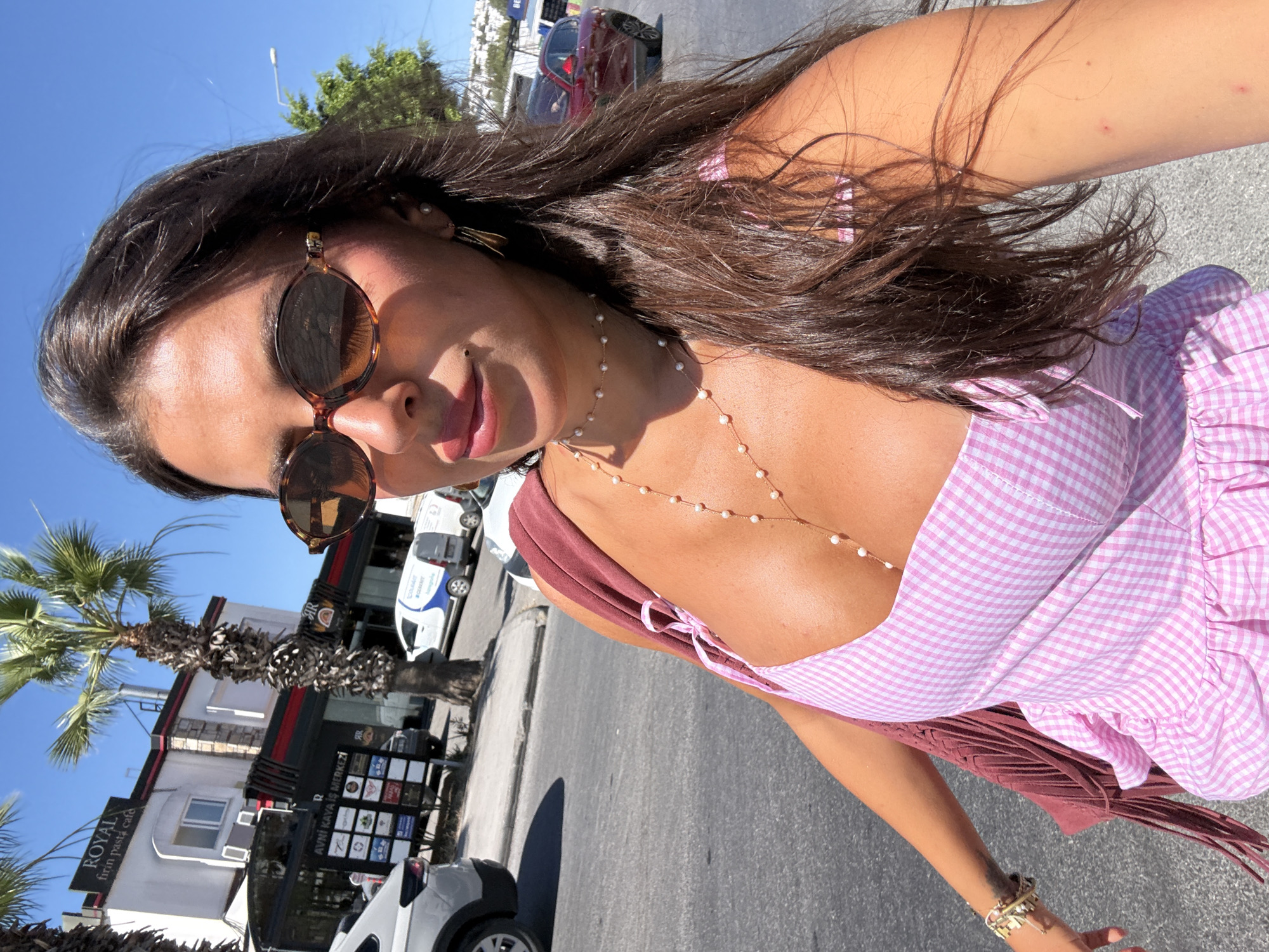 A woman with dark brown hair, wearing sunglasses and a pink gingham dress, takes a selfie outdoors in Bodrum, Turkey. The background features a white building with a "ROYAL fırın pasta cafe" sign, a palm tree, parked cars, and a street. She has a fringed bag slung over her shoulder and is wearing a pearl necklace and bracelets. The sun is shining brightly, casting shadows on the pavement.