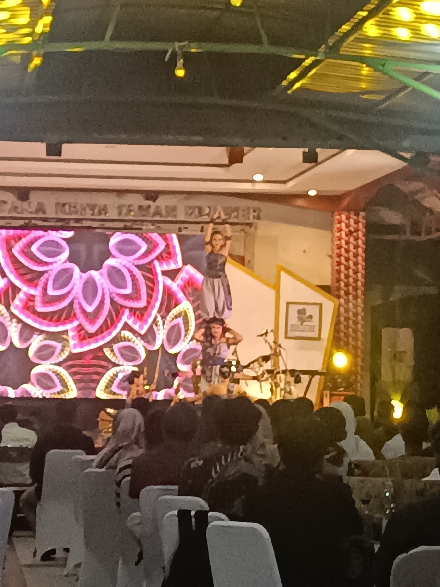 The image captures an outdoor performance at night, likely in Condongcatur, Indonesia, under a covered structure with yellow lighting visible on the ceiling. A large screen displays a vibrant pink and purple floral pattern. On stage, two individuals are performing. One woman, wearing a grey and purple outfit, stands on a raised platform with her arms outstretched. Below her, another person, also in a costume, is positioned near musical equipment and drums. In the foreground, an audience is seated on chairs, facing the stage. The audience members are largely obscured by the darkness and the lighting of the stage. The overall atmosphere suggests a cultural event or performance.