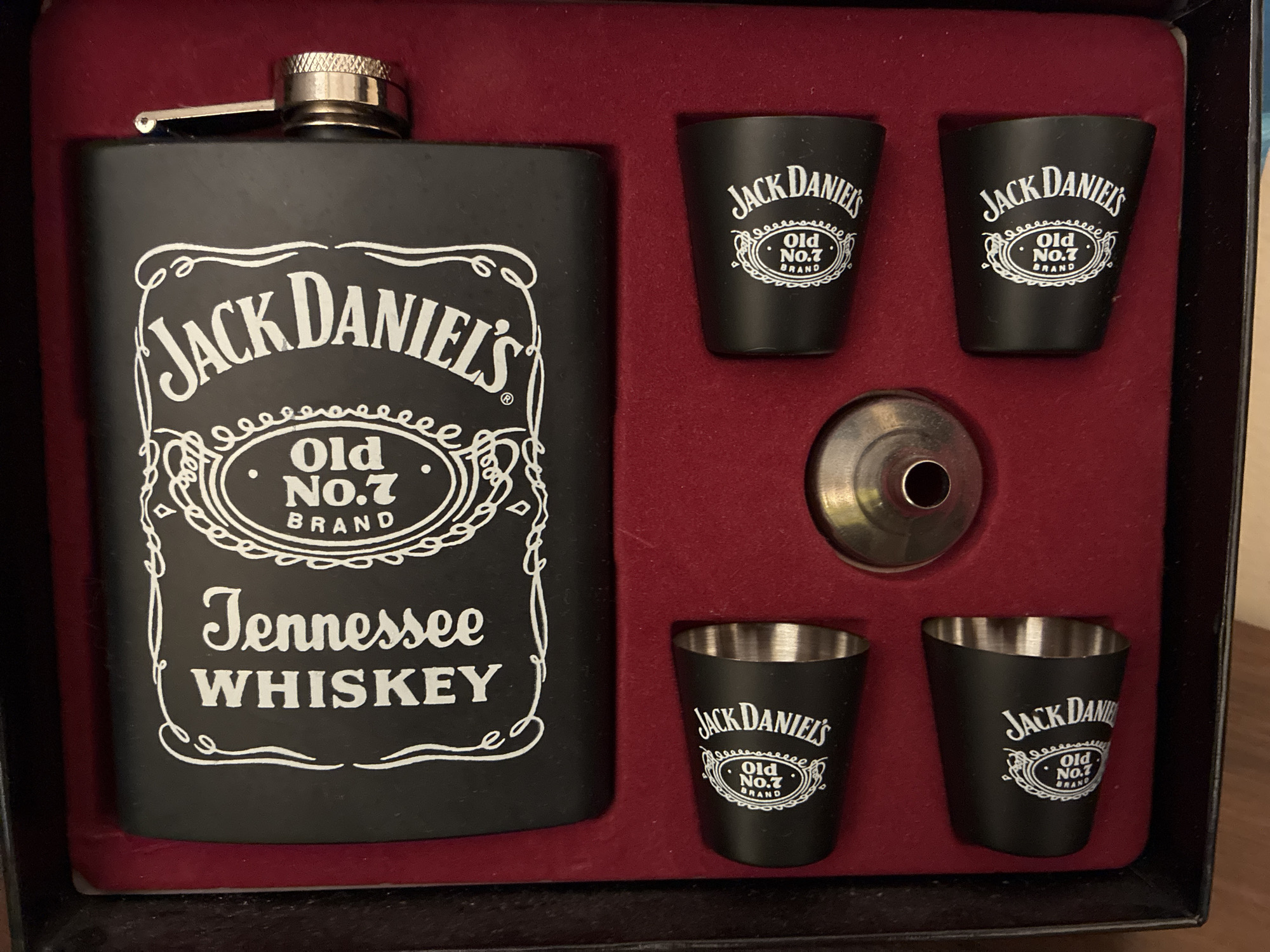 The image displays a Jack Daniel's branded gift set, neatly arranged within a red velvet-lined box. The set comprises a matte black hip flask, four matching shot glasses, and a small metallic funnel.
The hip flask, situated on the left, features the prominent Jack Daniel's label in white. Visible text on the flask includes "JACK DANIEL'S", "Old No.7 BRAND" (encased in an ornate oval border), and "Tennessee WHISKEY". The flask is topped with a silver-colored, textured screw cap, which is secured by a small metallic arm.
To the right, four small shot glasses are individually cradled in the red lining. These glasses are matte black with metallic interiors, each bearing the white "JACK DANIEL'S" logo and "Old No.7 BRAND" in the same decorative oval design.
In the center, between the upper and lower pairs of shot glasses, a small, polished metallic funnel rests in a circular cut-out. The overall presentation suggests a high-quality product or gift item, with the deep red interior of the box providing a striking contrast to the black and silver components of the set. The items appear new and unused, designed for convenient storage or gifting.
