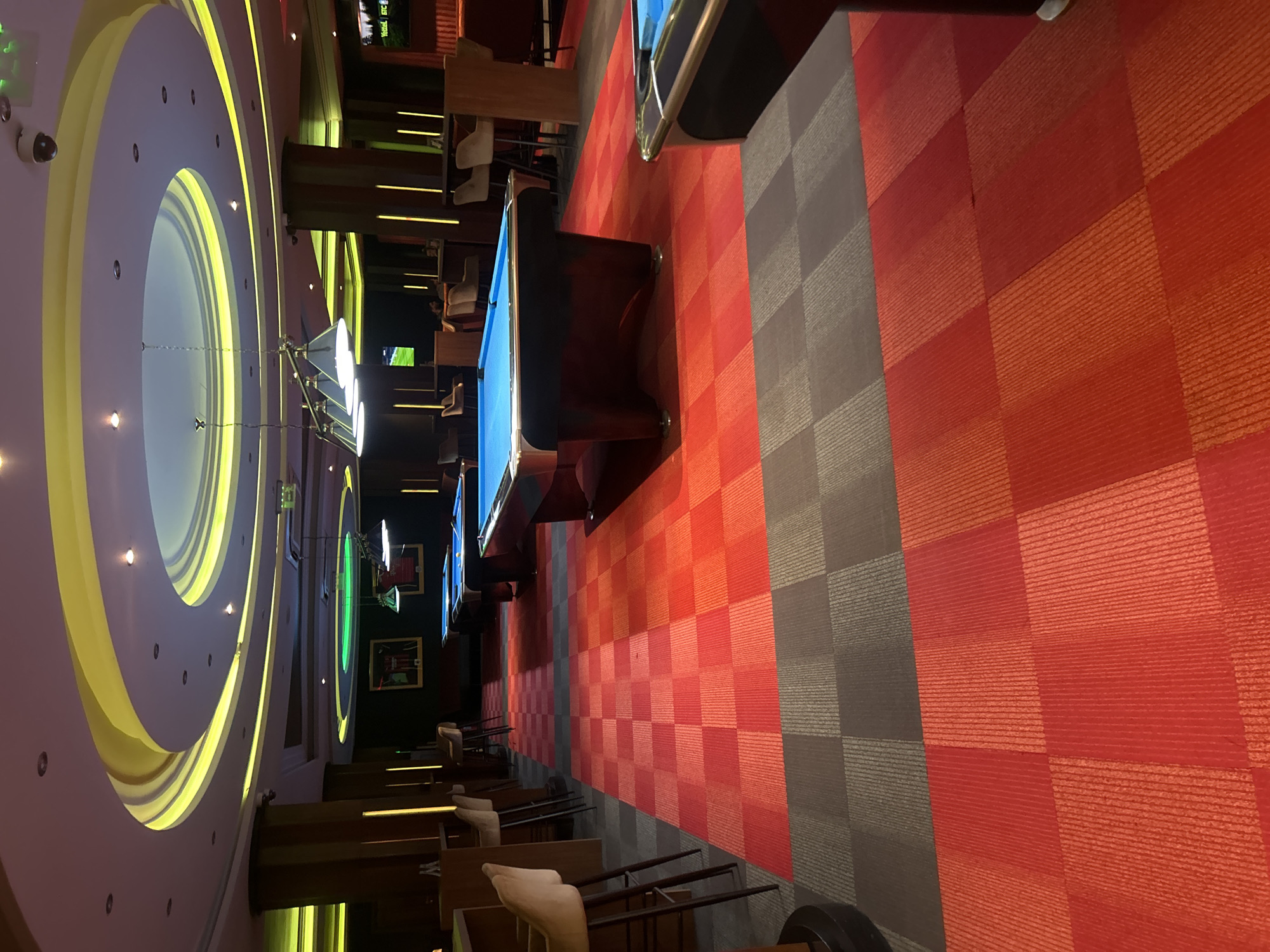 The image shows the interior of a recreational space, likely a bar or entertainment center. Several pool tables are visible, arranged in a row, with blue felt playing surfaces. Adjacent to the pool tables are bar stools, suggesting a casual seating arrangement for patrons. The flooring consists of alternating red and gray checkered tiles, creating a patterned design. The ceiling features a large, circular, layered lighting fixture with yellow illumination. The location is presumed to be in Sofia, Bulgaria. There is no evidence of people in the image.